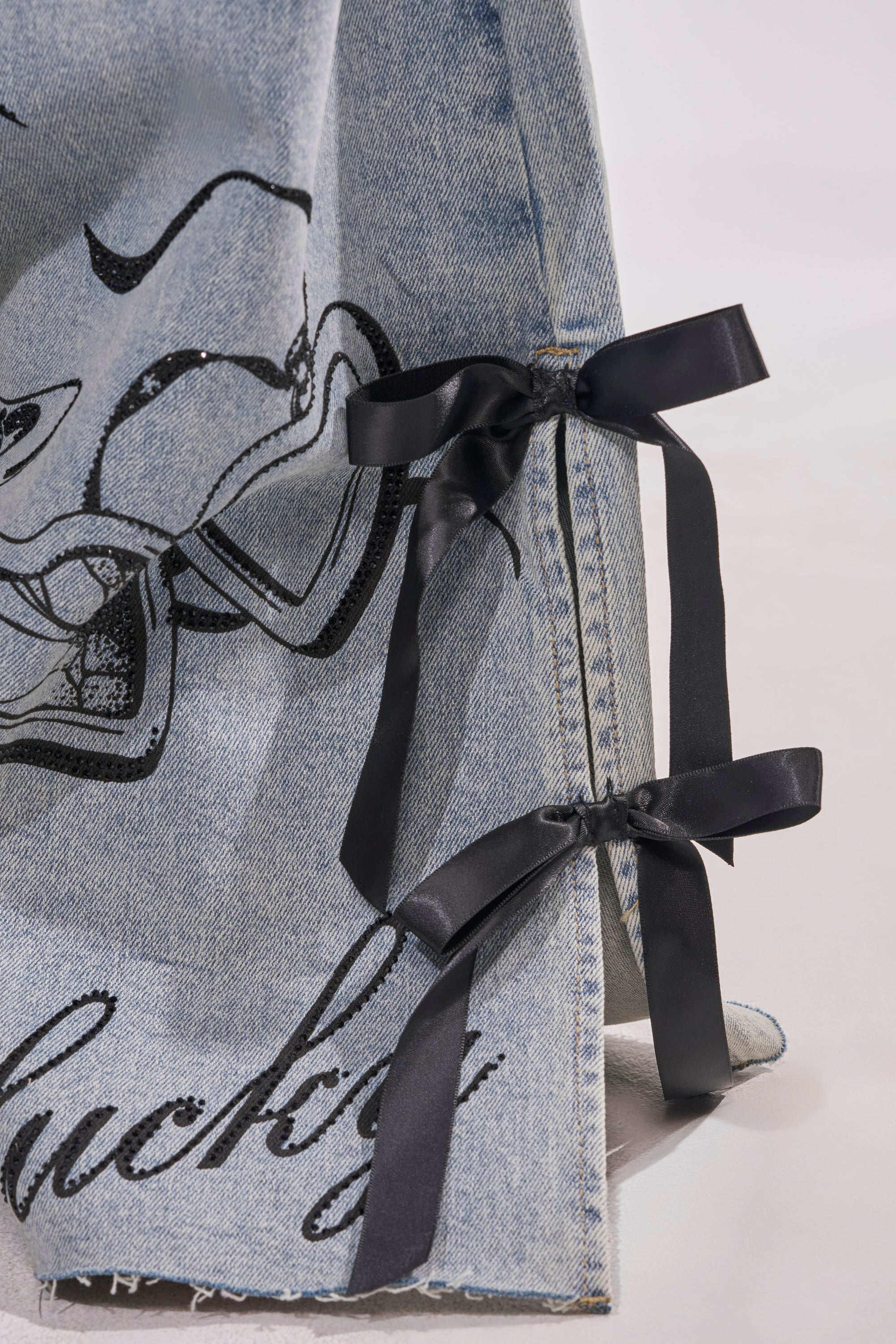 Close-up of the AFTER HOURS STRAIGHT LEG DENIM: light-wash jeans with black satin ribbon bows on the side seams, black printed graphics, and partial "lucky" text near the frayed hem. 8