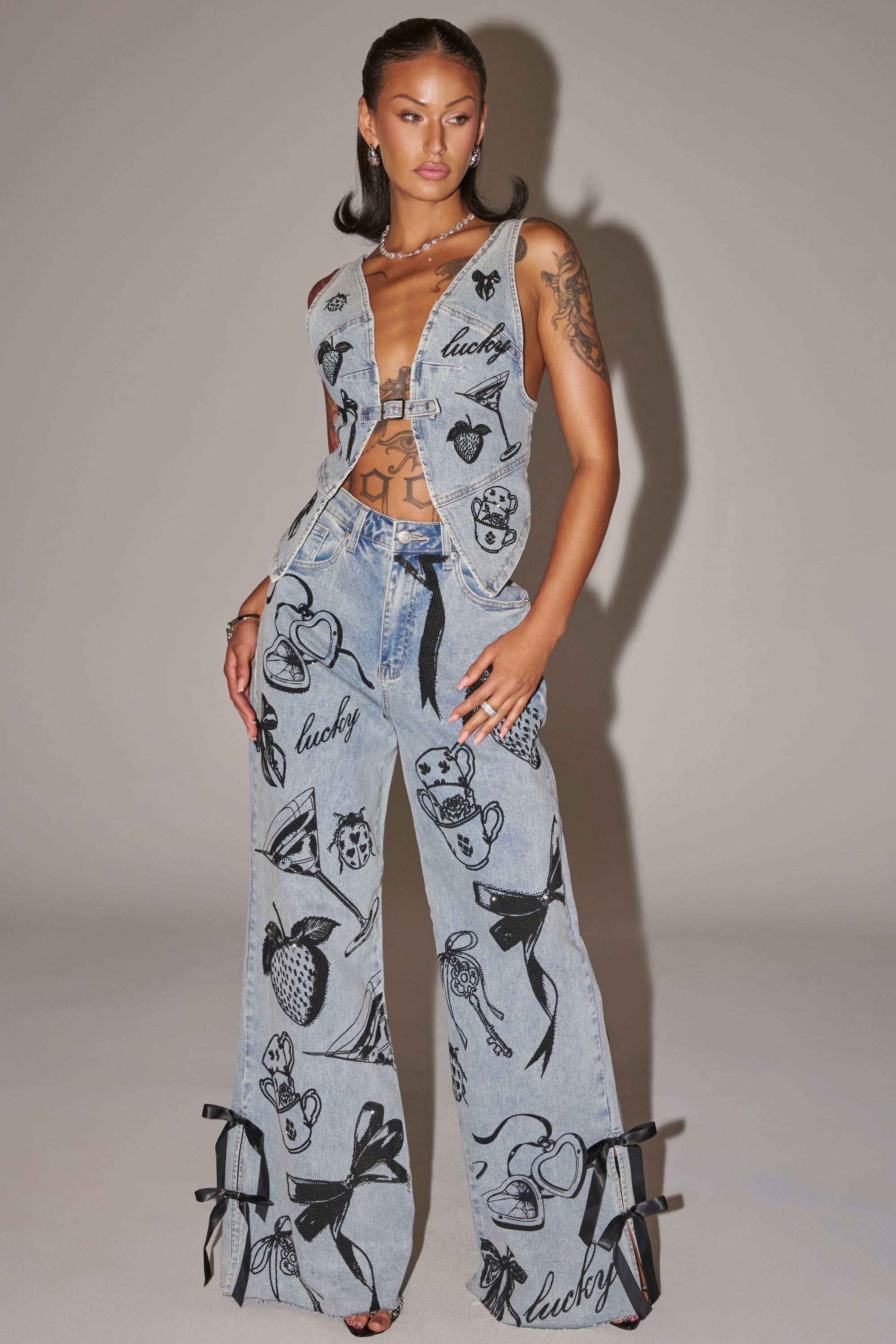 A woman wears the AFTER HOURS DENIM TOP, a cropped denim piece featuring bold black tattoo-inspired designs, bows, and "lucky" text. She pairs it with wide-leg jeans, jewelry, and shows off tattoos on her arm and torso. 5