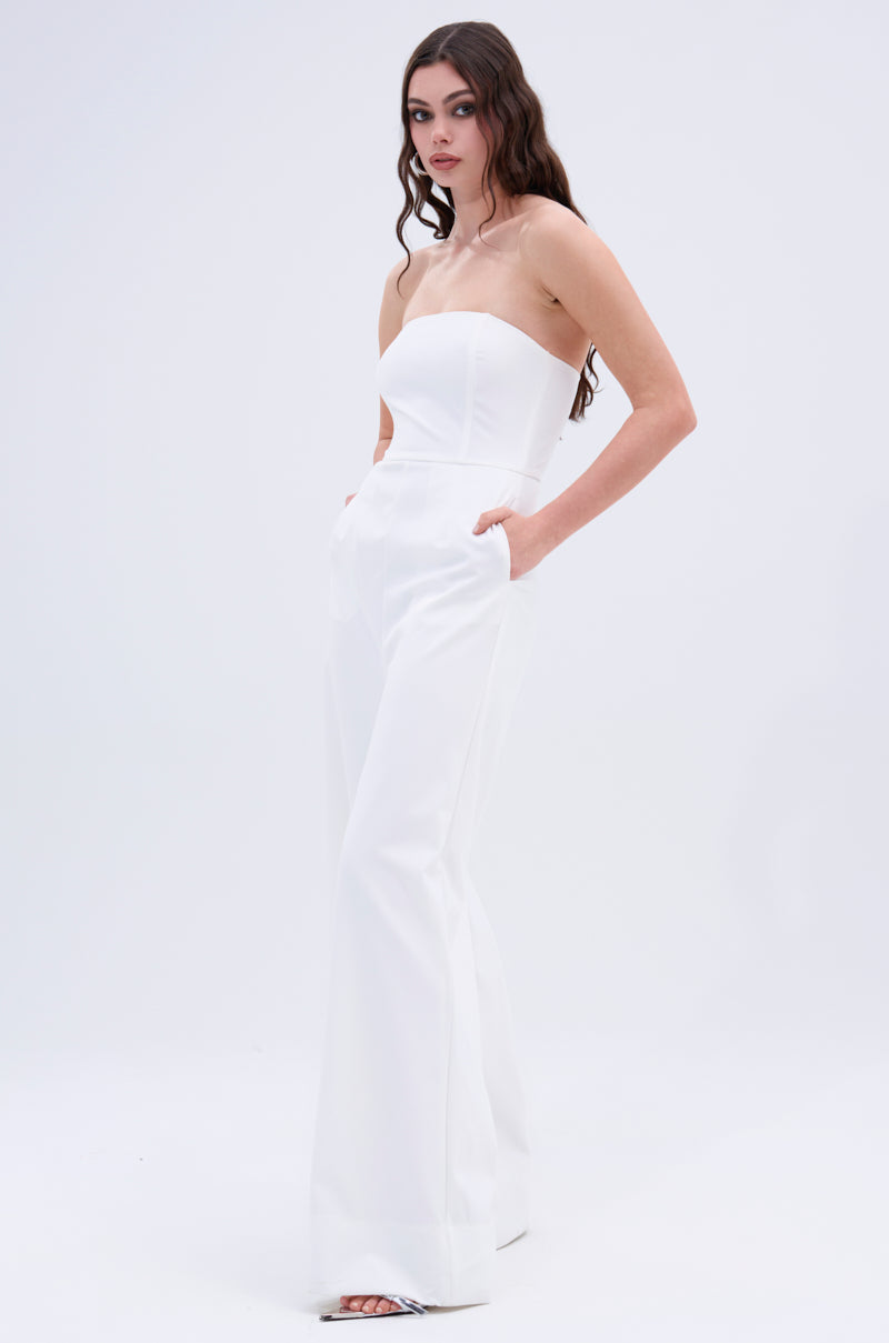 A woman wears the BIG BOSS ENERGY WIDE LEG JUMPSUIT IN WHITE, featuring a strapless neckline and metallic back zipper, standing against a white background with hands in pockets and long wavy hair over her shoulders.