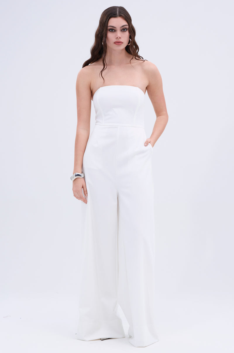 A woman with long, wavy brown hair exudes big boss energy in the BIG BOSS ENERGY WIDE LEG JUMPSUIT IN WHITE, featuring a strapless neckline and wide-leg fit as she poses confidently against a plain white background.