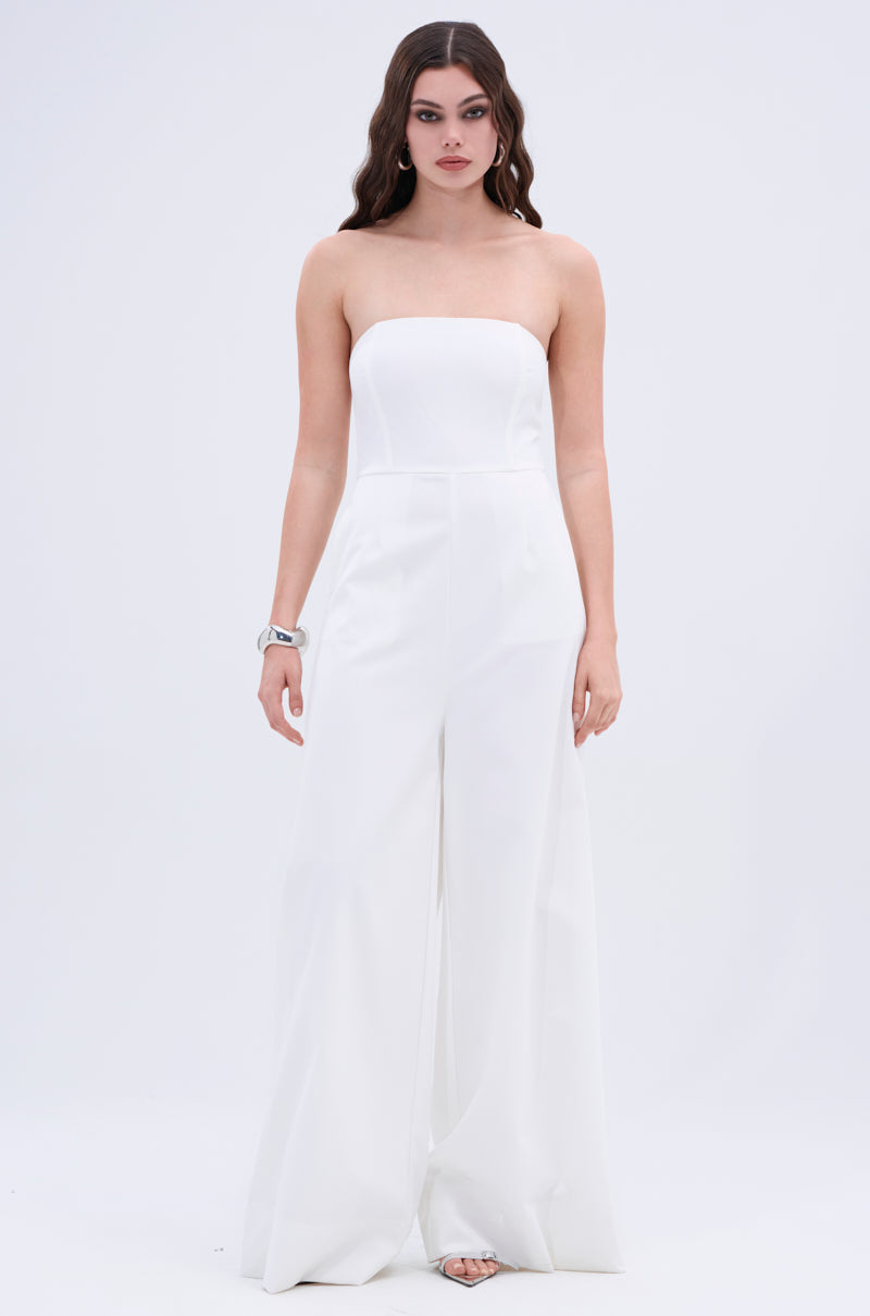 A woman with long wavy brown hair wears the BIG BOSS ENERGY WIDE LEG JUMPSUIT IN WHITE with open-toe heels and a silver bracelet, posing against a plain white background with a neutral expression.
