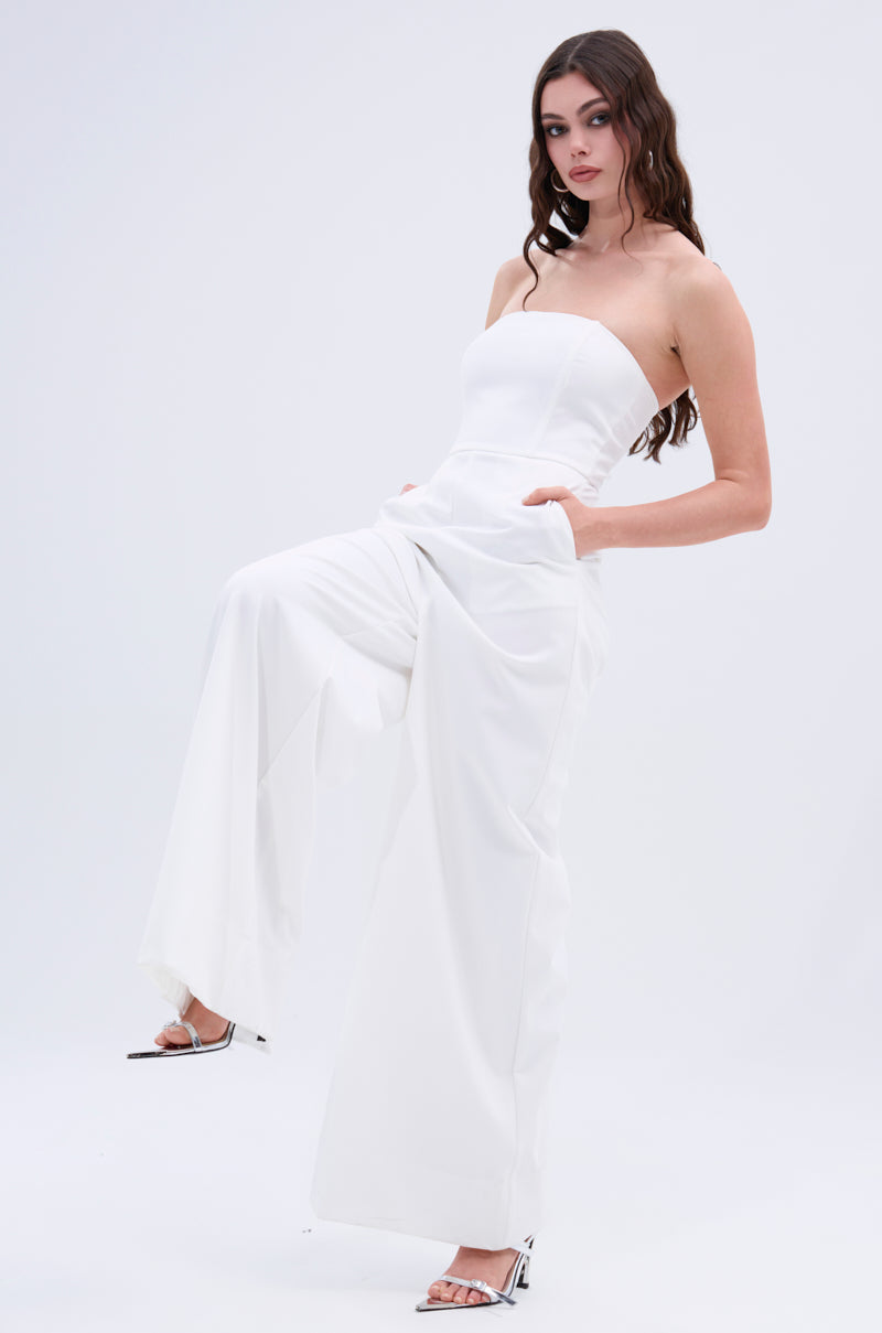 A woman poses against a plain light background in the BIG BOSS ENERGY WIDE LEG JUMPSUIT IN WHITE, styled with white heels, one leg raised and hands in her pockets.