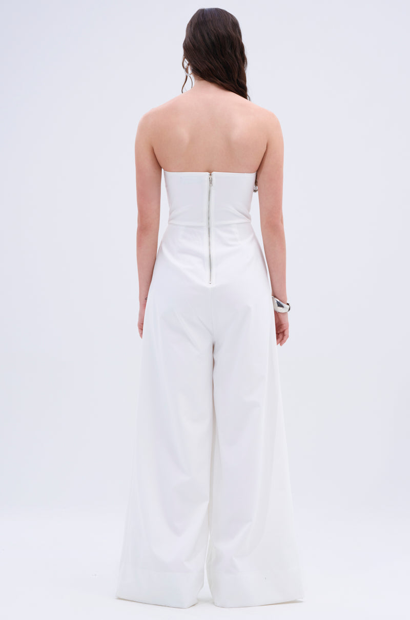 A woman faces away in the BIG BOSS ENERGY WIDE LEG JUMPSUIT IN WHITE, featuring a strapless neckline and metallic back zipper. She wears her long, dark hair loose and accessorizes with a chunky right-hand bracelet against a plain white background.