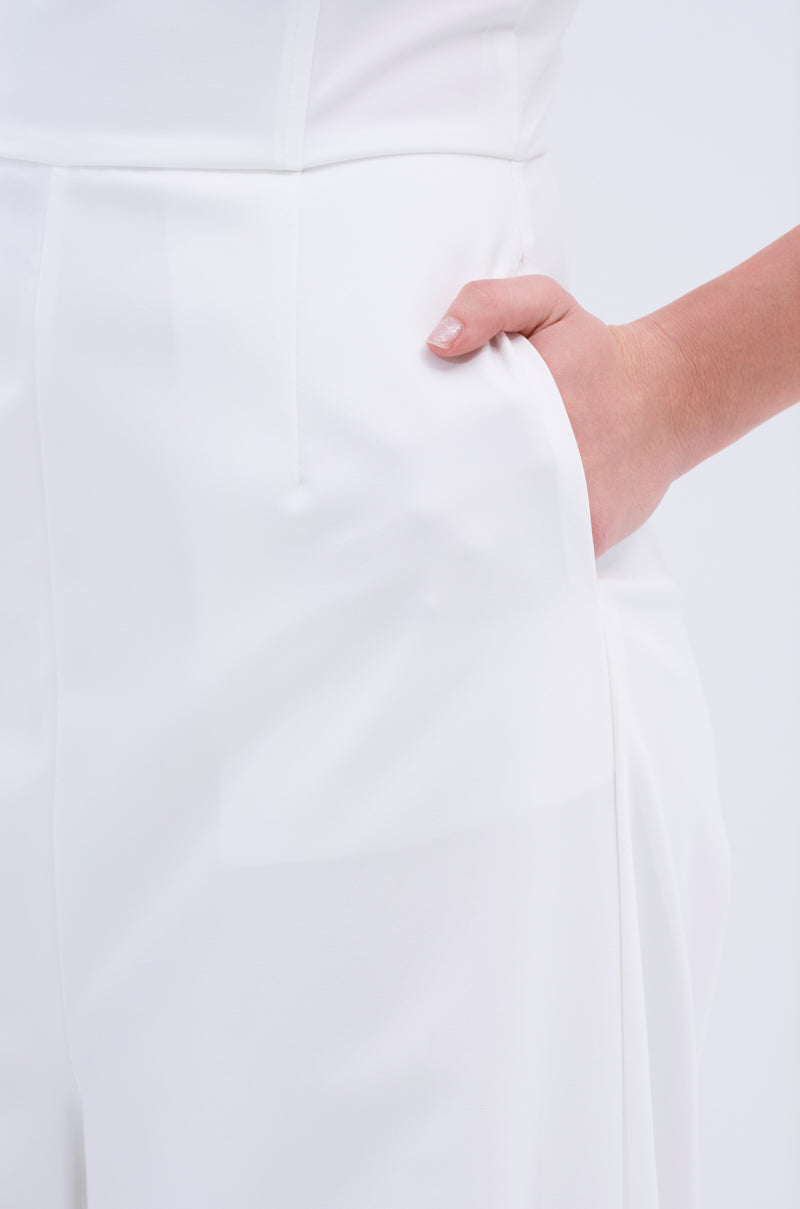 Close-up of a person wearing the BIG BOSS ENERGY WIDE LEG JUMPSUIT IN WHITE with their right hand in a side pocket, against a plain, light background that highlights its minimalist, modern silhouette.