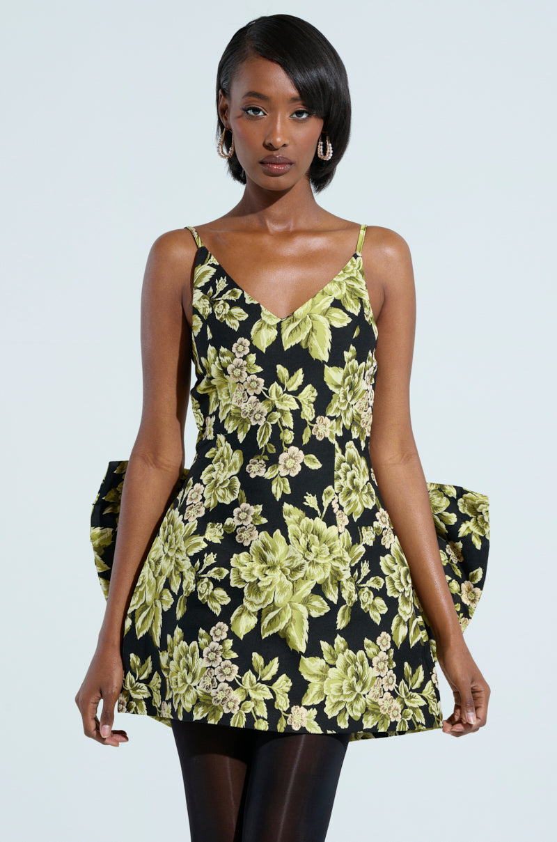 A person is wearing the "Business in the Back Brocade Mini Dress with Bow," featuring a black base, green floral pattern, thin straps, a short hemline, and a large bow at the back. They have shoulder-length hair and are looking straight ahead against a plain, light-colored background.