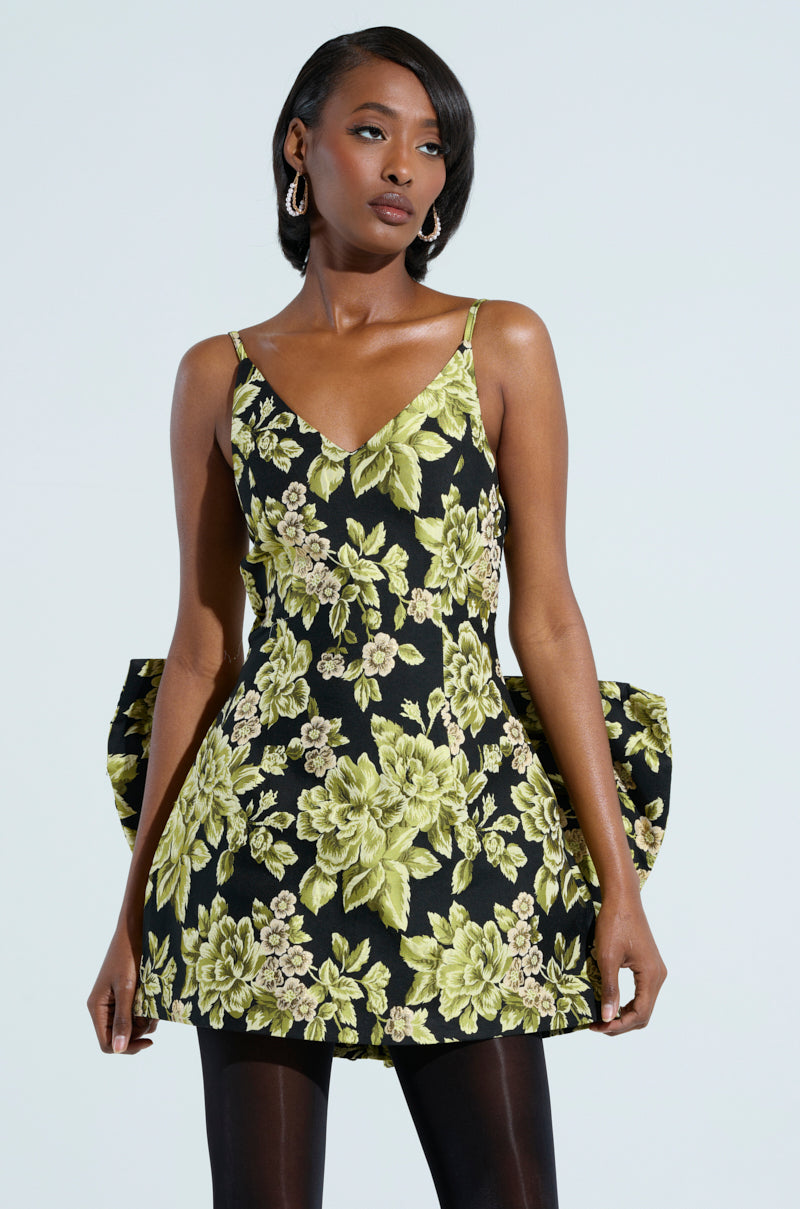 Dressed in the BUSINESS IN THE BACK BROCADE MINI DRESS WITH BOW, a person stands against a light background. The black dress features bold green floral patterns and a back bow. With shoulder-length dark hair and hoop earrings, they confidently gaze to the side.