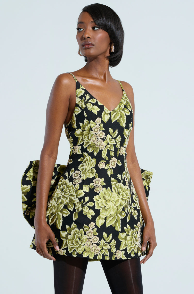A woman poses in the BUSINESS IN THE BACK BROCADE MINI DRESS WITH BOW, flaunting a black floral pattern with large green and cream flowers. The design is highlighted by a deep V-neckline, thin straps, and a large ruffle at the back. Her sleek side-parted black hair complements the outfit perfectly.