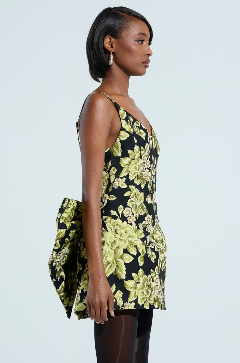 A woman in profile against a light background wears the "Business in the Back" Brocade Mini Dress, featuring a sleeveless, party-perfect floral pattern and a large bow on the back.