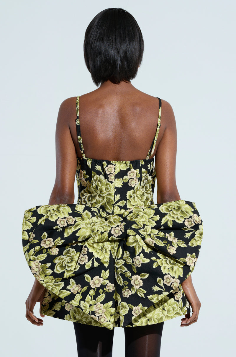 A person with short hair, seen from the back, wears the "Business in the Back Brocade Mini Dress With Bow," featuring a black and green floral pattern and thin straps. This party-perfect dress includes a decorative bow at the waist, against a light gray background.