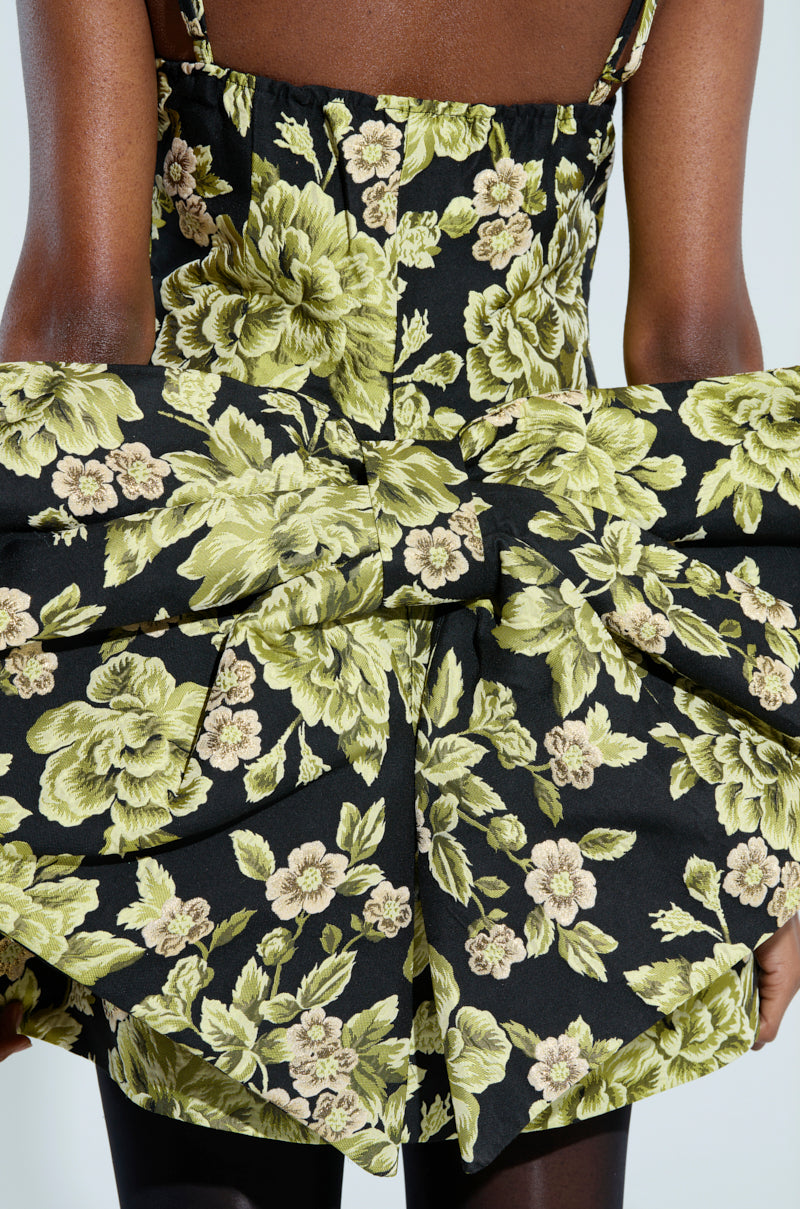 A person wears the "Business in the Back Brocade Mini Dress with Bow," showcasing a floral pattern with large green flowers on a black background. The dress features an elegant bow at the back, highlighting their arms and upper torso beautifully.