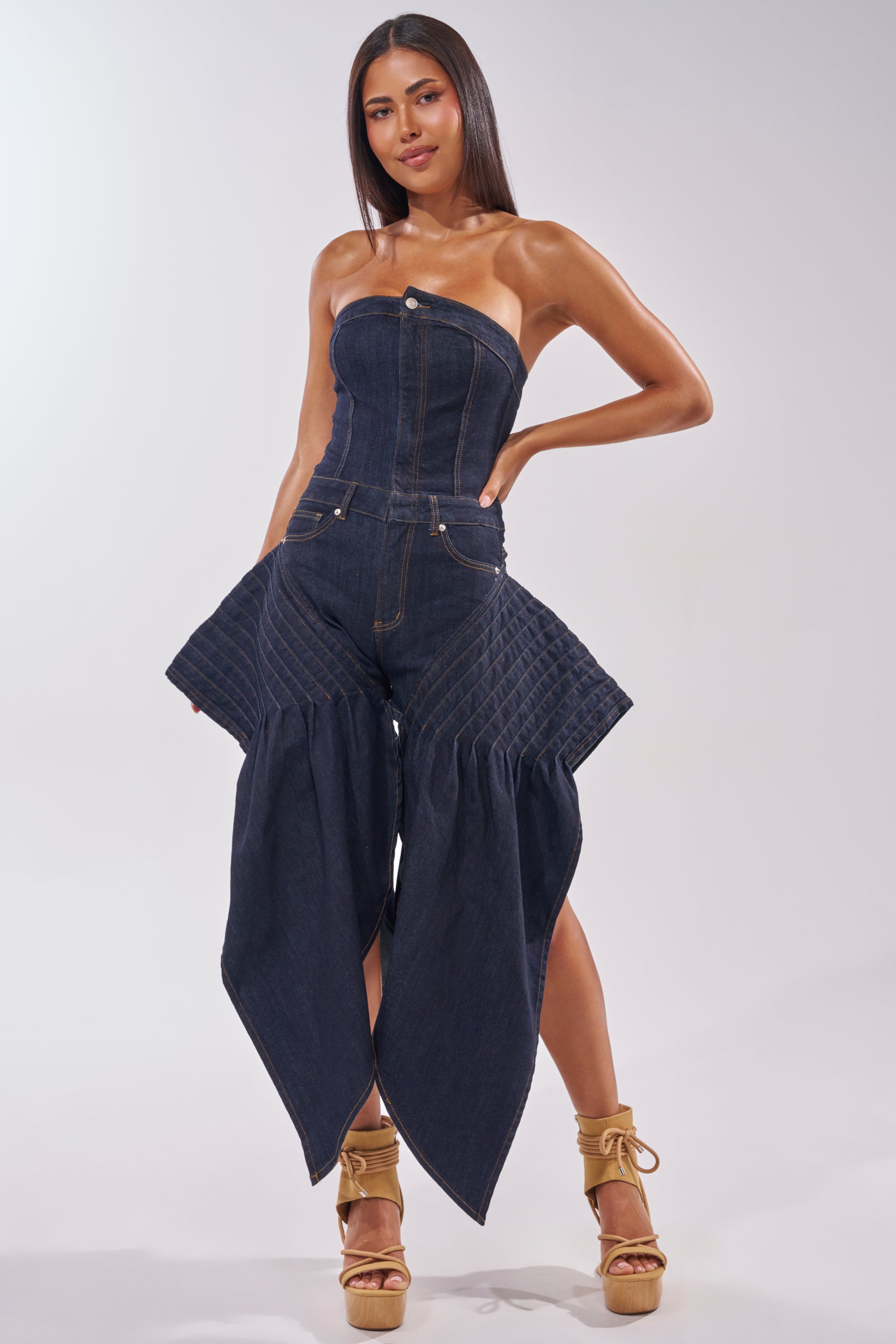 A woman poses confidently against a light background wearing the NO ONE LIKE ME DENIM ROMPER IN DARK BLUE DENIM, a strapless jumpsuit with exaggerated hip detailing and flared panels. She pairs it with tan platform heels and has long, straight hair. 0
