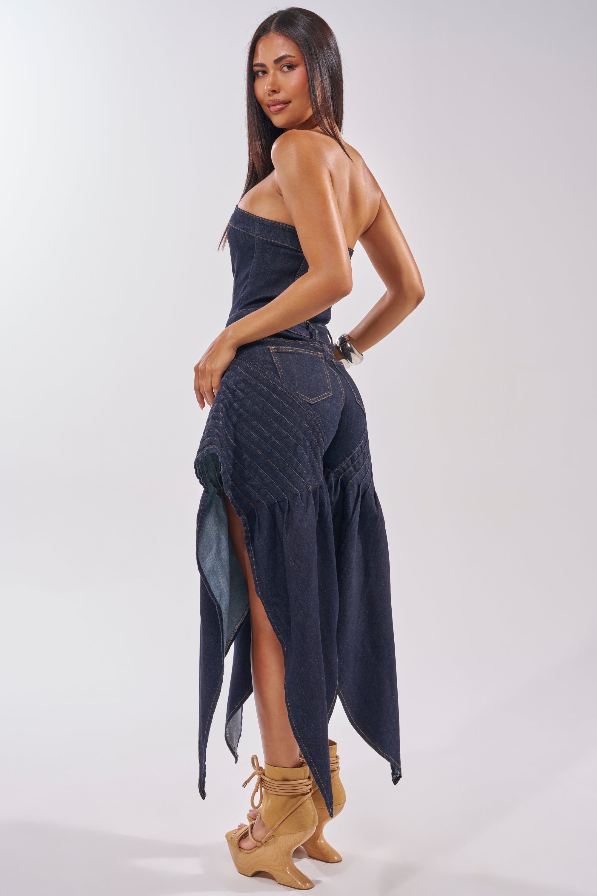 A woman with long straight hair poses in the NO ONE LIKE ME DENIM ROMPER IN DARK BLUE DENIM, which features a strapless corset bodice and high slits. She pairs it with tan lace-up heeled sandals, looking over her shoulder against a light backdrop. 1