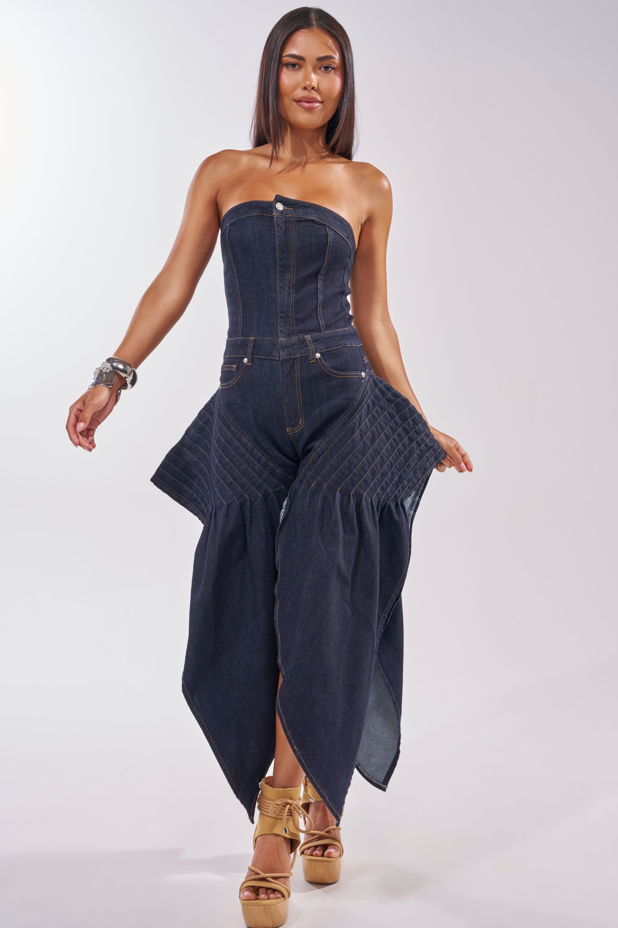 A woman stands confidently in the NO ONE LIKE ME DENIM ROMPER IN DARK BLUE DENIM, a strapless, form-fitting piece with a corset bodice and statement hips, styled with gold platform sandals and silver bangles against a plain light background. 2