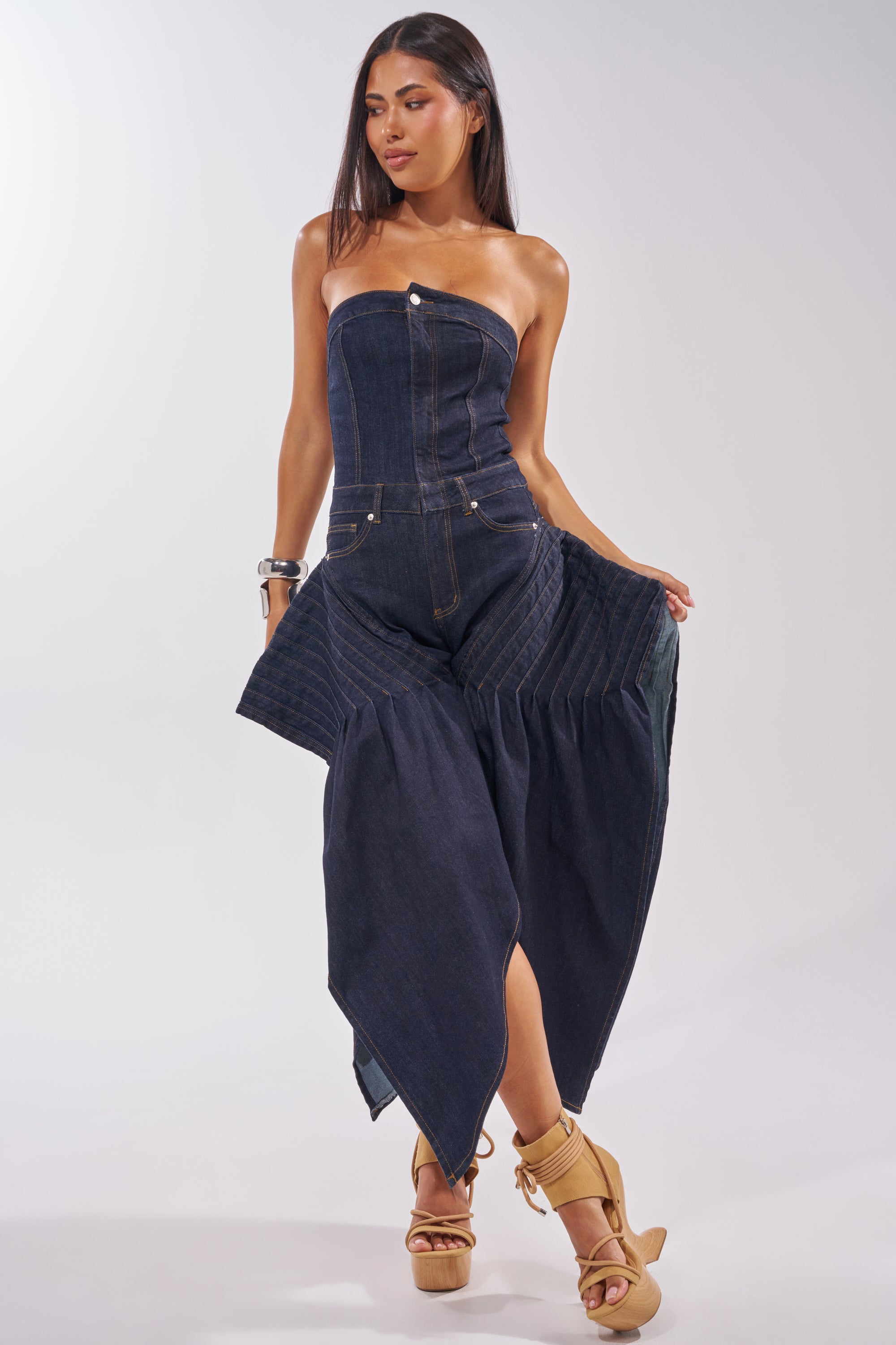 A woman wearing the NO ONE LIKE ME DENIM ROMPER IN DARK BLUE DENIM, featuring a corset-style bodice and flared asymmetrical skirt, stands against a light background in tan platform heels while holding her skirt. 3