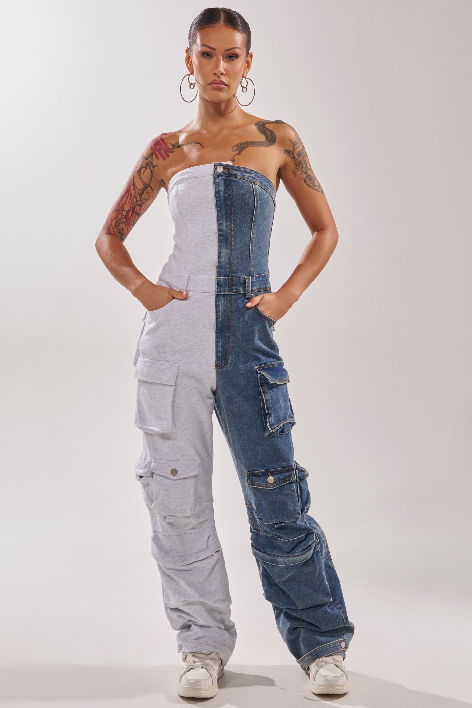 A woman exudes confidence in the SPLIT DECISION DENIM JUMPSUIT—a strapless jumpsuit with cargo pockets, featuring one side gray fabric and the other blue denim. She completes the look with sneakers, hoop earrings, and visible tattoos. 2