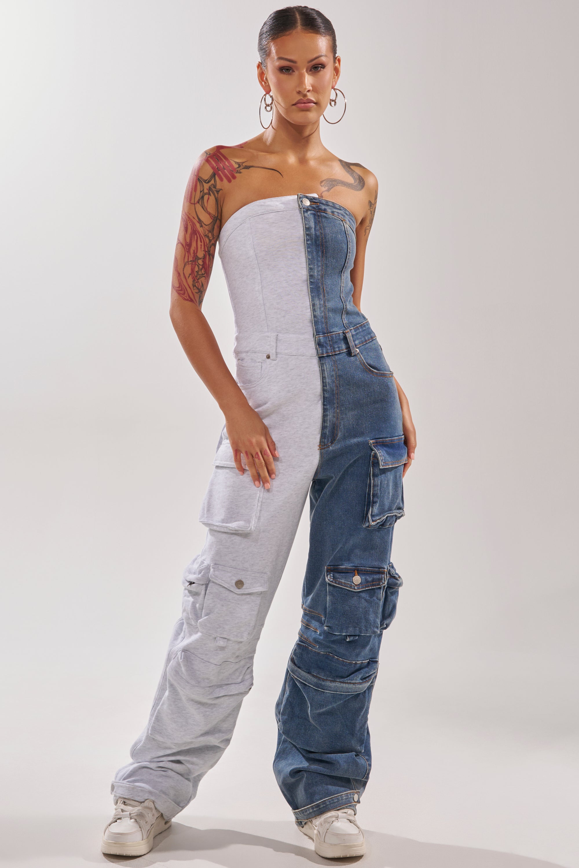 A person exudes confidence in the SPLIT DECISION DENIM JUMPSUIT, a strapless one-piece with gray sweatshirt fabric on one side, blue denim on the other, and large cargo pockets. Hoop earrings, visible tattoos, and white sneakers complete the bold look. 3