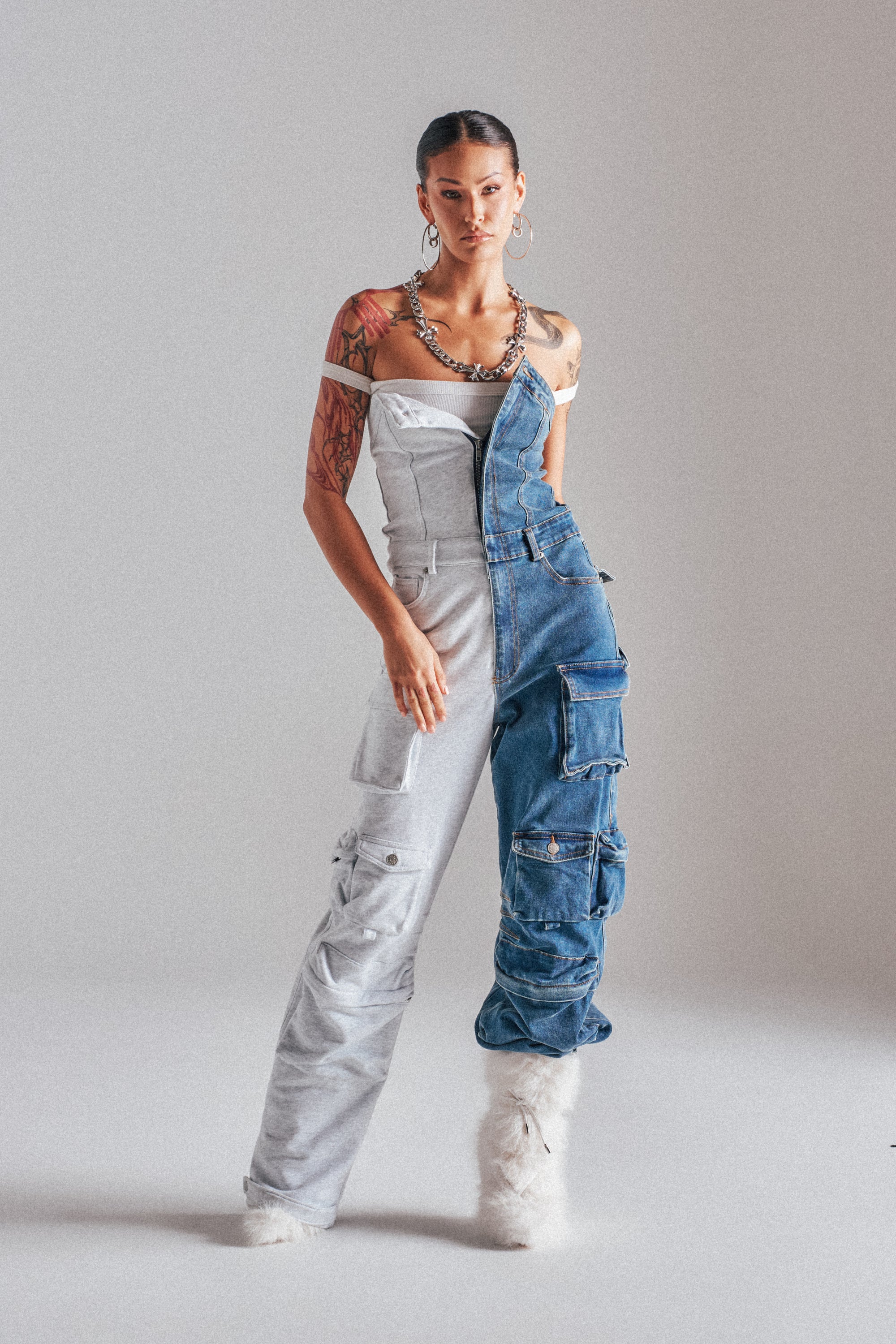 A model in the studio wears the SPLIT DECISION DENIM JUMPSUIT, a strapless, asymmetrical piece with half light gray fabric and half blue denim, featuring multiple cargo pockets. Their look is styled with tattoos, hoop earrings, a chain necklace, and white furry shoes. 1