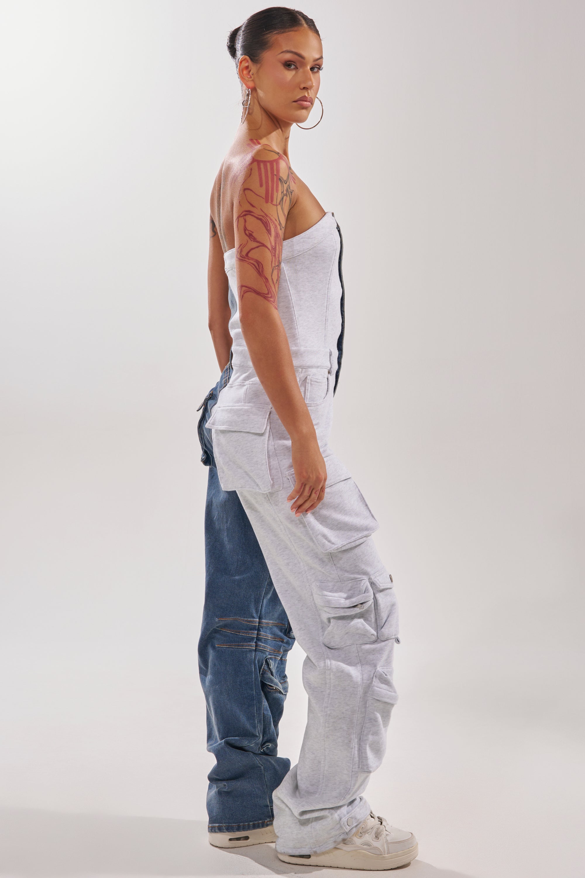 A woman in profile wears the SPLIT DECISION DENIM JUMPSUIT—half denim, half gray cargo sweatpant style—paired with a white strapless top, hoop earrings, a large arm tattoo, and white sneakers. 4