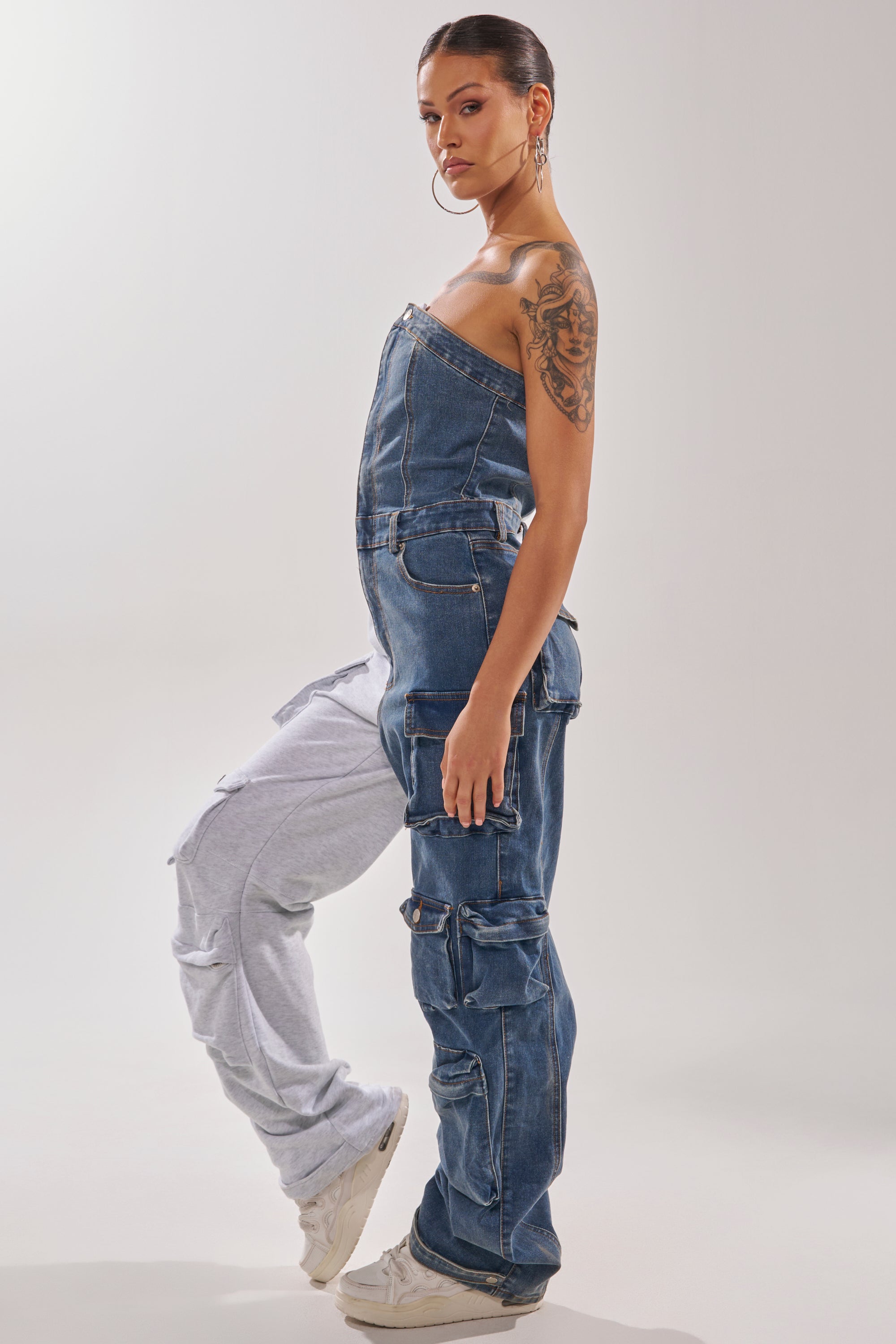 A woman models the SPLIT DECISION DENIM JUMPSUIT—half denim with cargo pockets, half gray sweatpants—paired with white sneakers and hoop earrings, showing an arm tattoo against a plain backdrop. 5