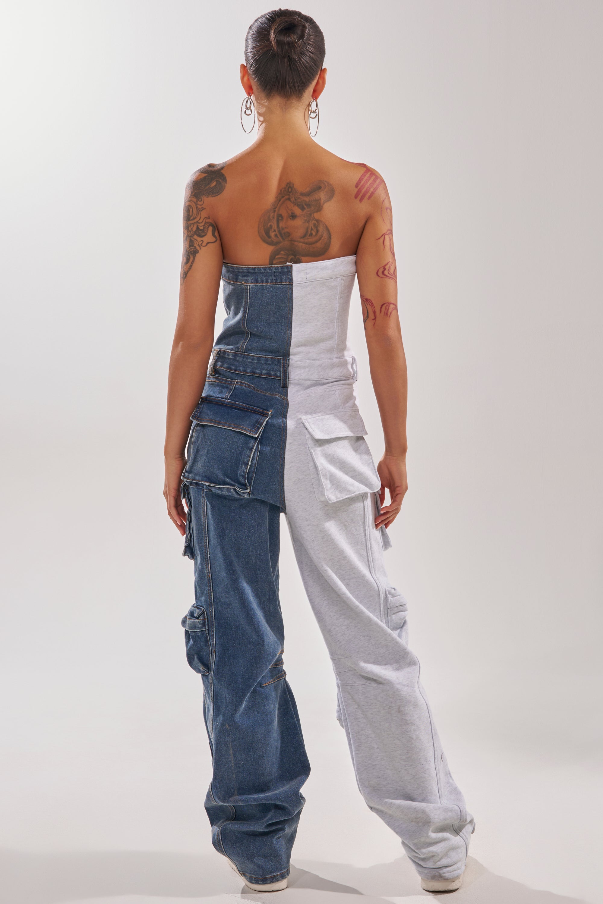 A person stands facing away in the SPLIT DECISION DENIM JUMPSUIT—strapless, with blue denim on one side and gray sweatshirt fabric on the other, cargo pockets, visible arm and back tattoos, and hair in a bun. 6