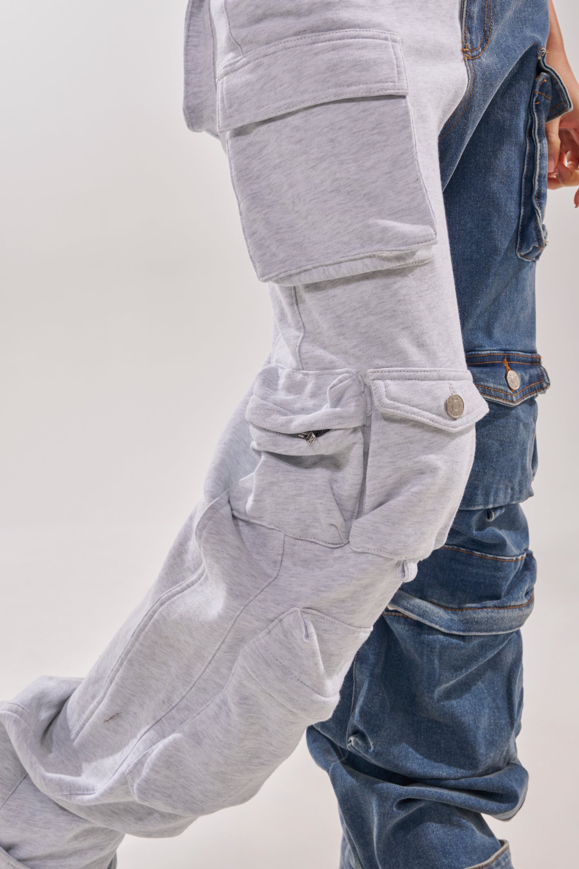 Close-up of a person wearing the SPLIT DECISION DENIM JUMPSUIT, featuring one grey sweatshirt leg and one blue denim leg with matching cargo pockets, standing against a plain background. 8