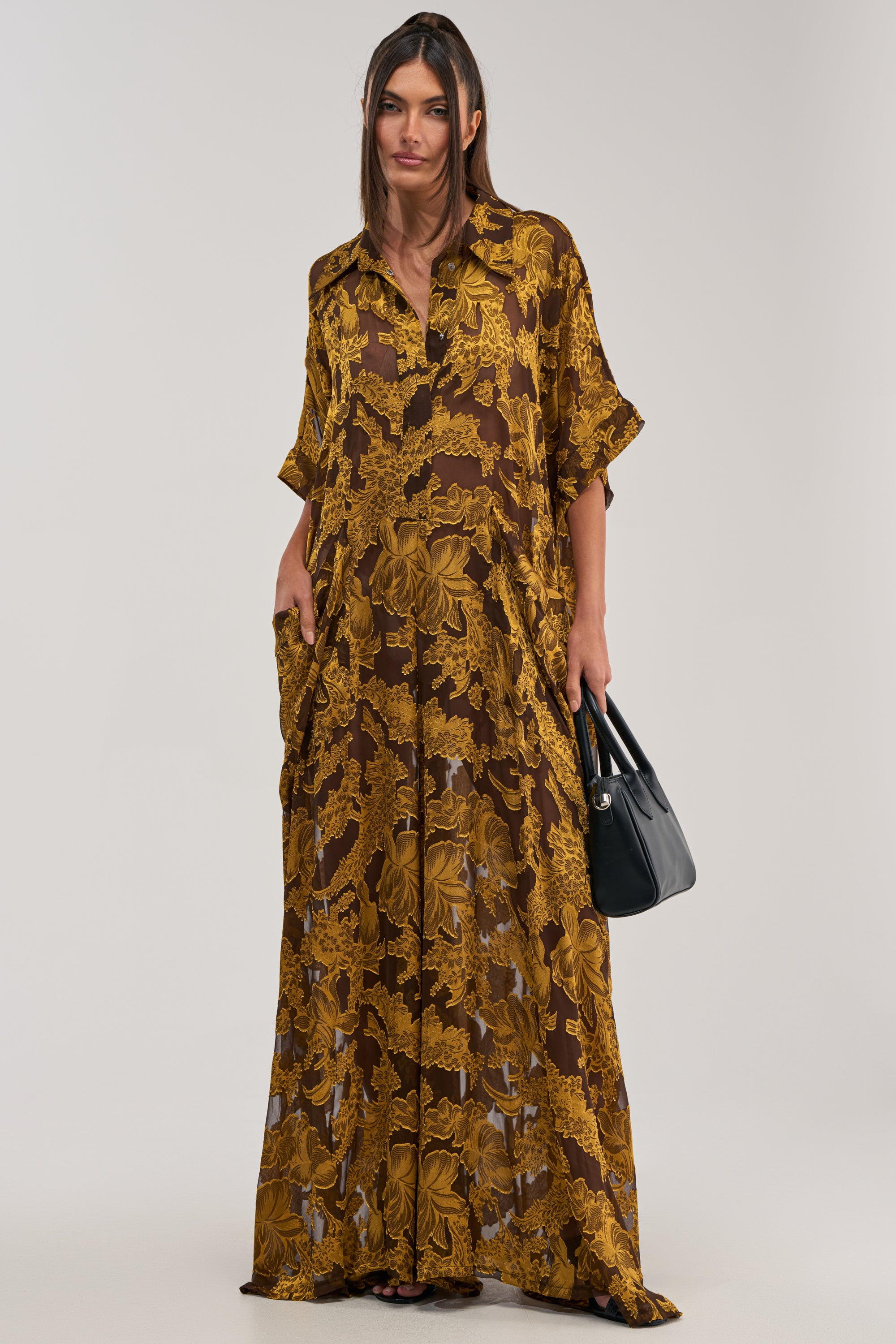 Wearing the DEEP IN THE FEELS LACE JUMPSUIT IN YELLOW MULTI, a woman with long brown hair stands holding a black handbag. The loose, floor-length jumpsuit features a yellow floral pattern against a simple, light background.