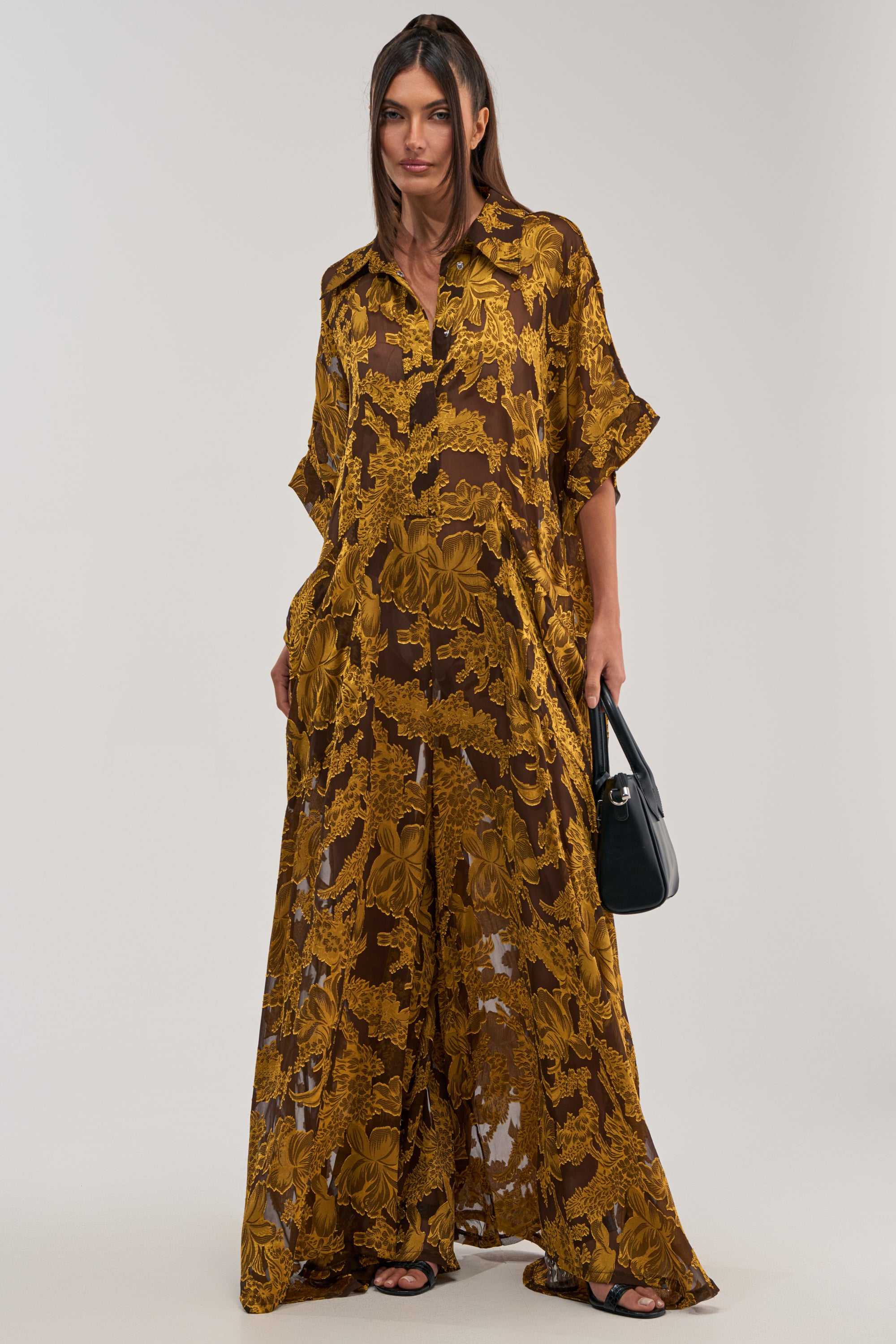 A woman wears the DEEP IN THE FEELS LACE JUMPSUIT IN YELLOW MULTI, featuring a loose fit and vibrant floral pattern with short sleeves and a collar. She pairs it with a black handbag and sandals, standing against a plain light background.