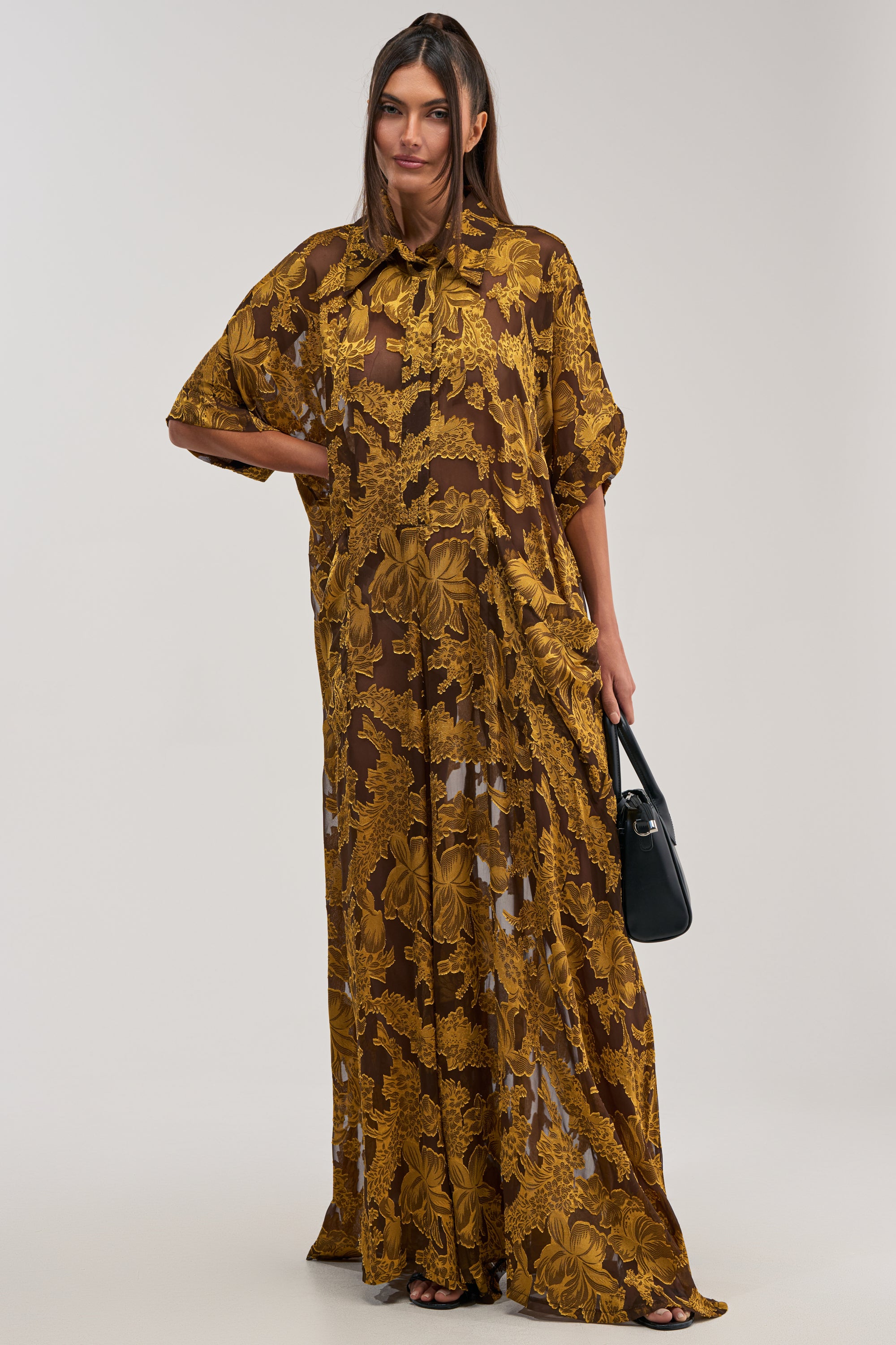 A woman stands against a plain background wearing the DEEP IN THE FEELS LACE JUMPSUIT IN YELLOW MULTI. She holds a black handbag, and her long straight hair complements the jumpsuit's vibrant boho-chic style.