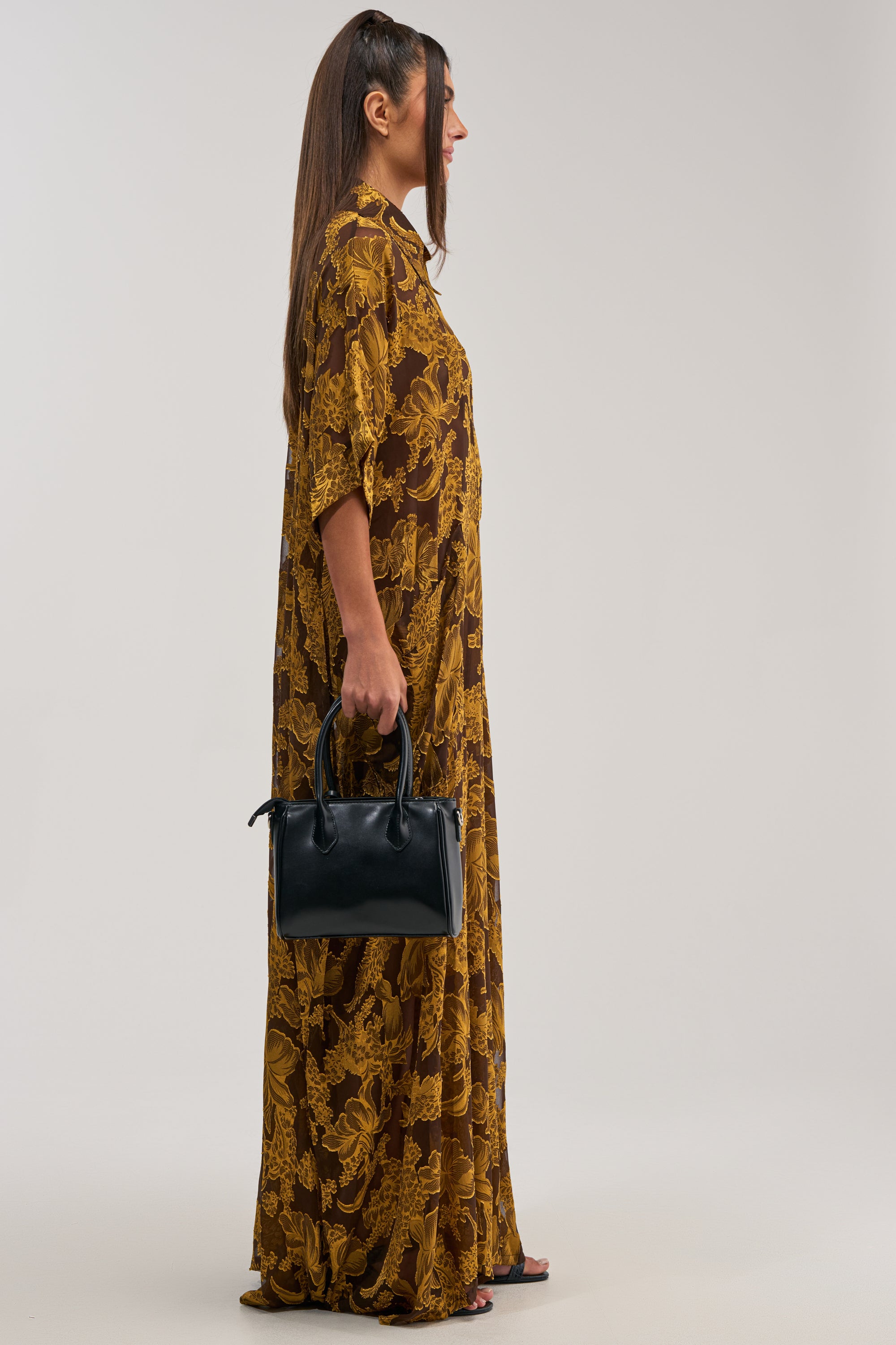 A woman stands in profile wearing the DEEP IN THE FEELS LACE JUMPSUIT IN YELLOW MULTI, holding a black handbag and wearing black sandals, set against a plain light background.