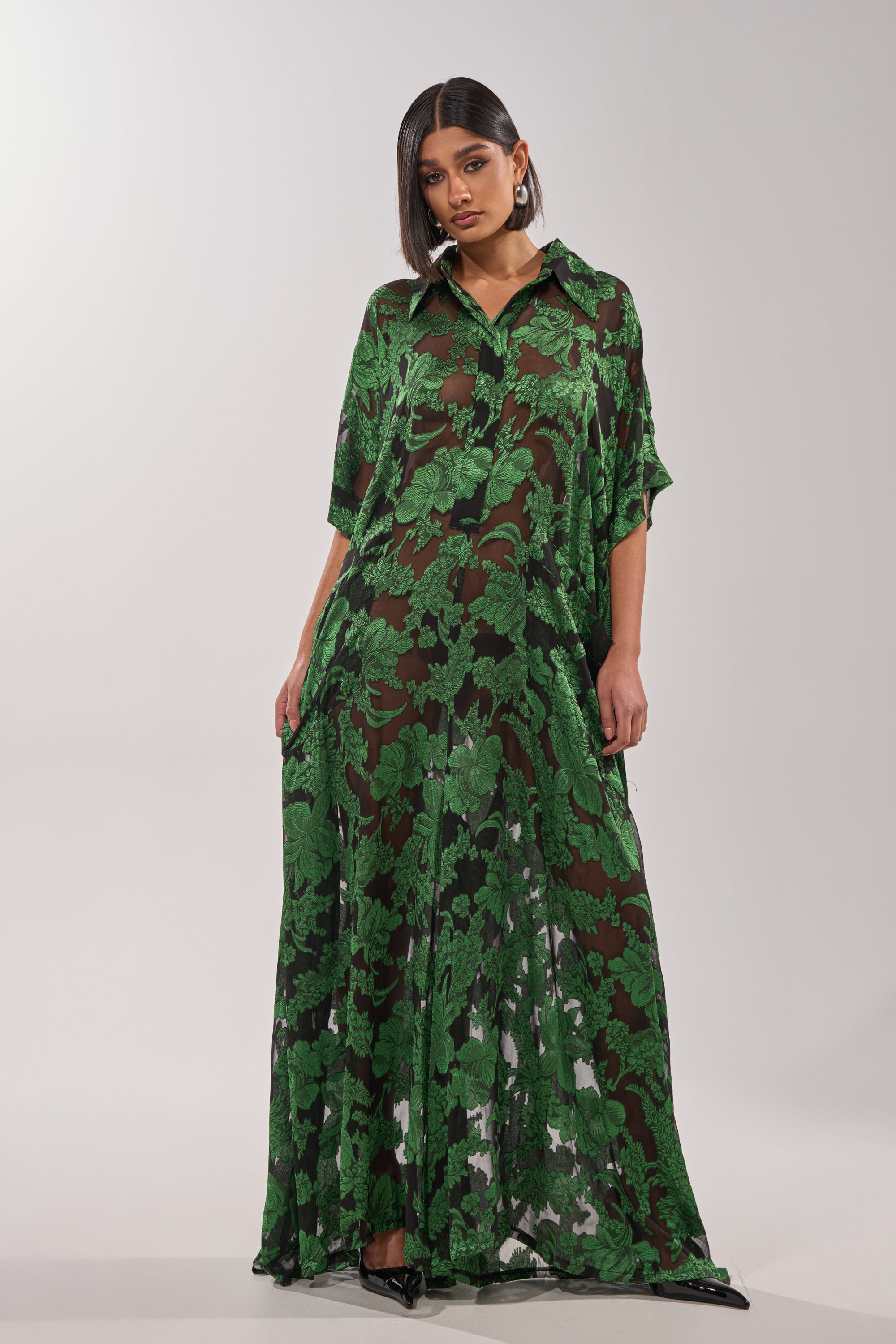 A woman models the DEEP IN THE FEELS LACE JUMPSUIT, a long, loose-fit botanical lace piece featuring a green leafy pattern on a dark base. She stands before a light backdrop, wearing statement earrings and pointed shoes with straight dark hair. 0