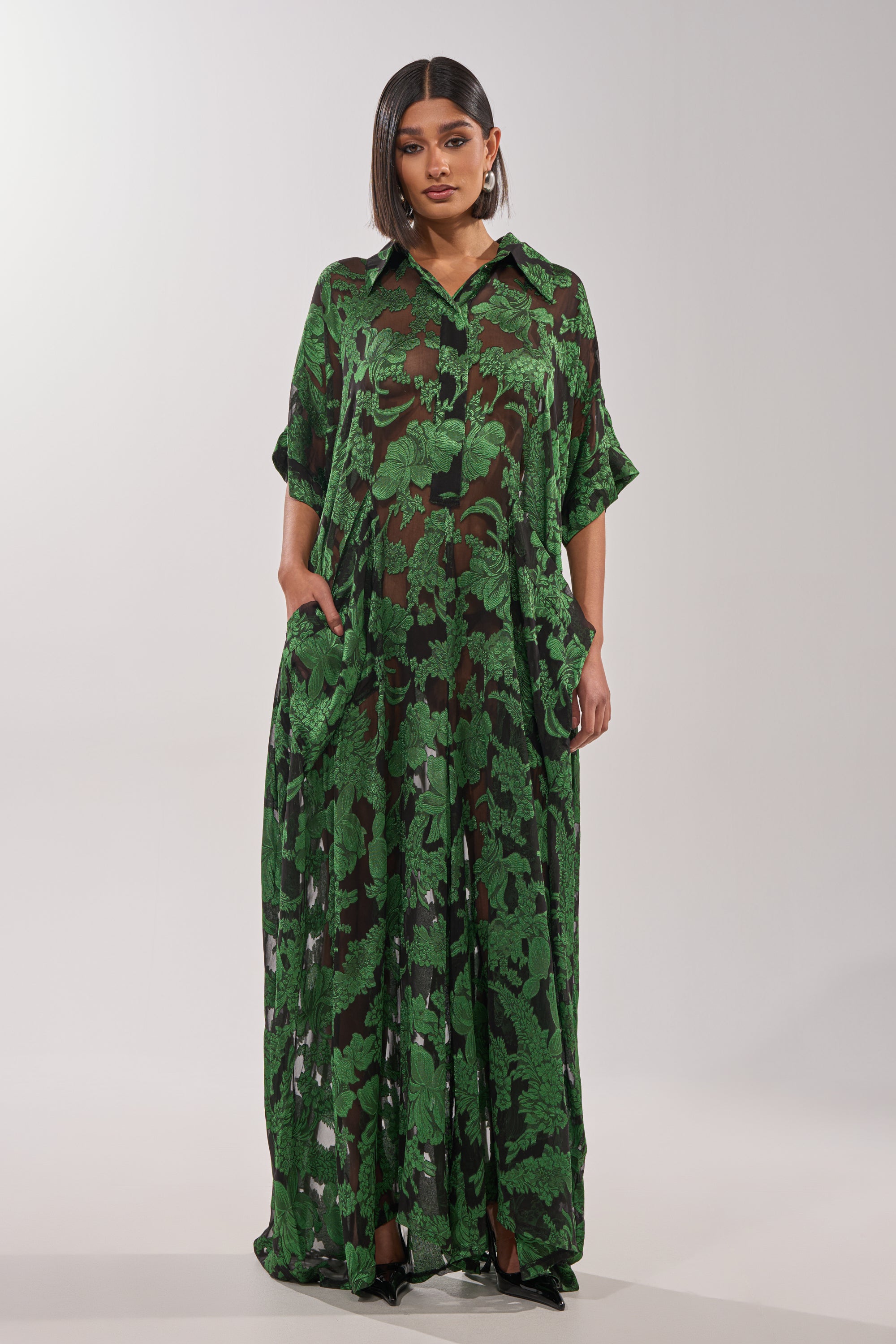 A woman with a sleek bob and neutral makeup stands facing forward in the DEEP IN THE FEELS LACE JUMPSUIT, featuring a sheer botanical leaf pattern, short sleeves, and pockets, set against a plain light background. 1
