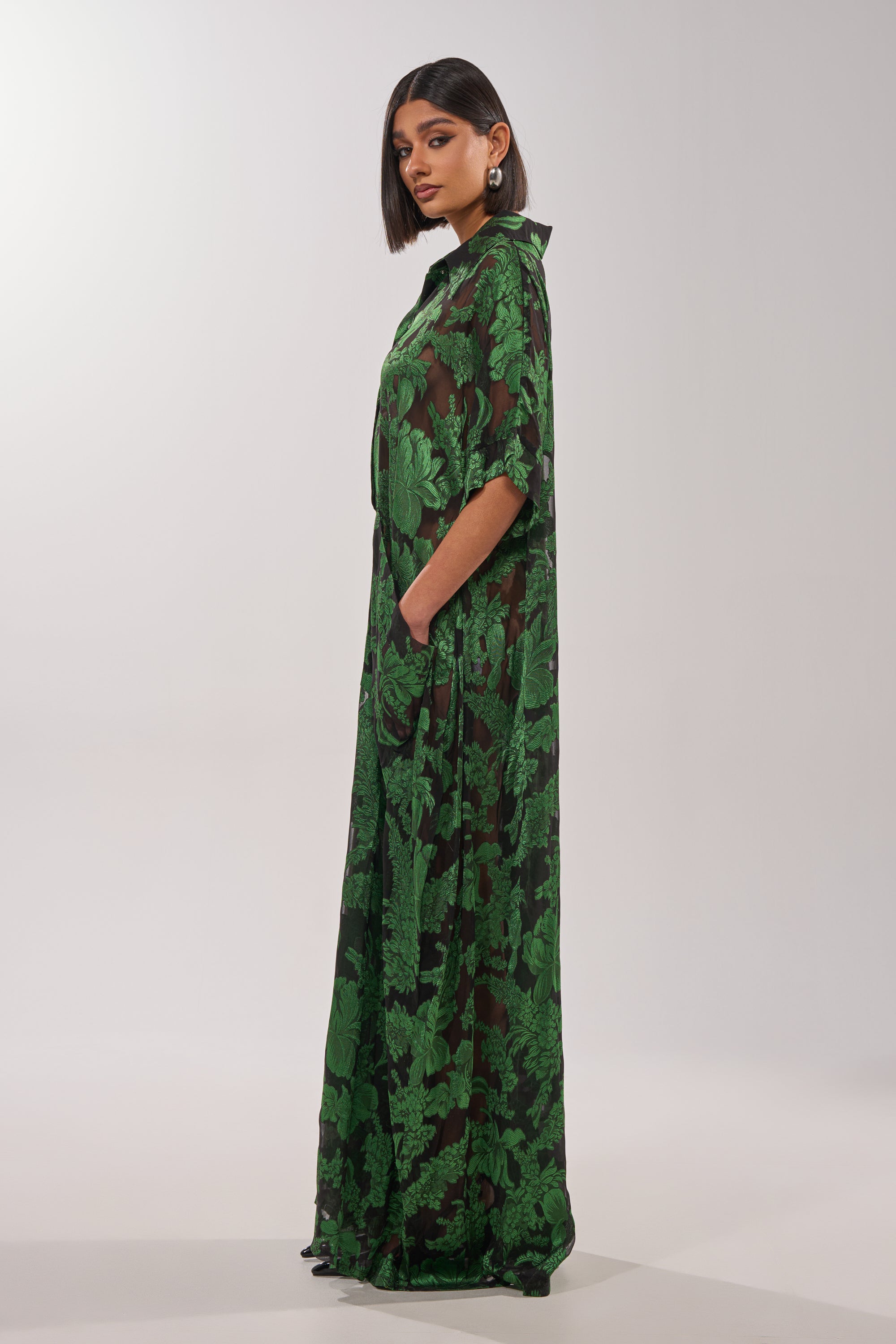 A woman with a bob poses sideways in the DEEP IN THE FEELS LACE JUMPSUIT—a long, sheer black piece with bold green leaf patterns—hands in her pockets, set against a plain white background. 3