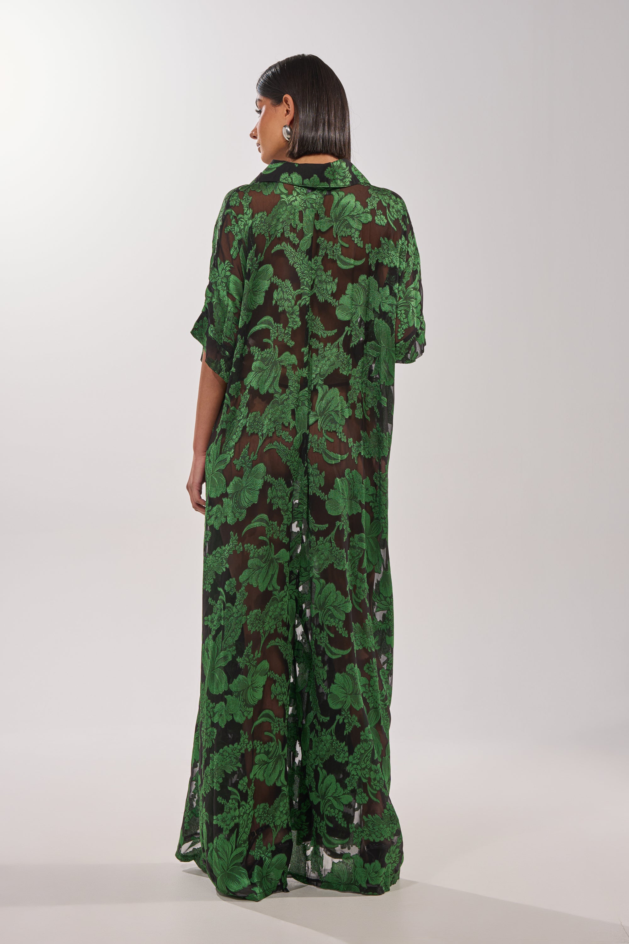 A person faces away wearing the DEEP IN THE FEELS LACE JUMPSUIT, a long, loose-fitting sheer outfit with a leafy lace pattern, short sleeves, and set against a plain light background. 4