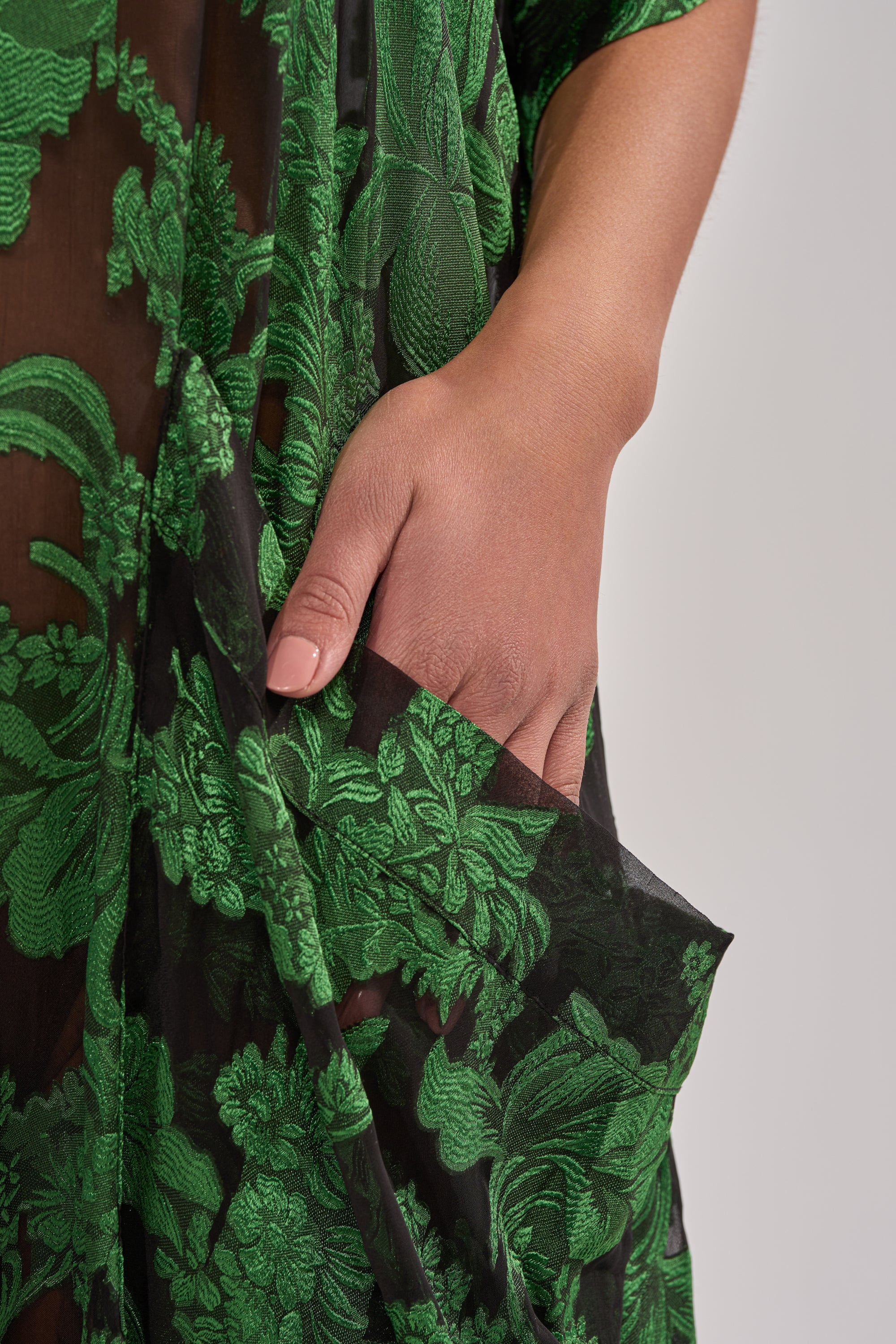 A close-up of a hand resting in the pocket of the DEEP IN THE FEELS LACE JUMPSUIT, featuring sheer black botanical lace and green floral embroidery, against a plain light background. 5