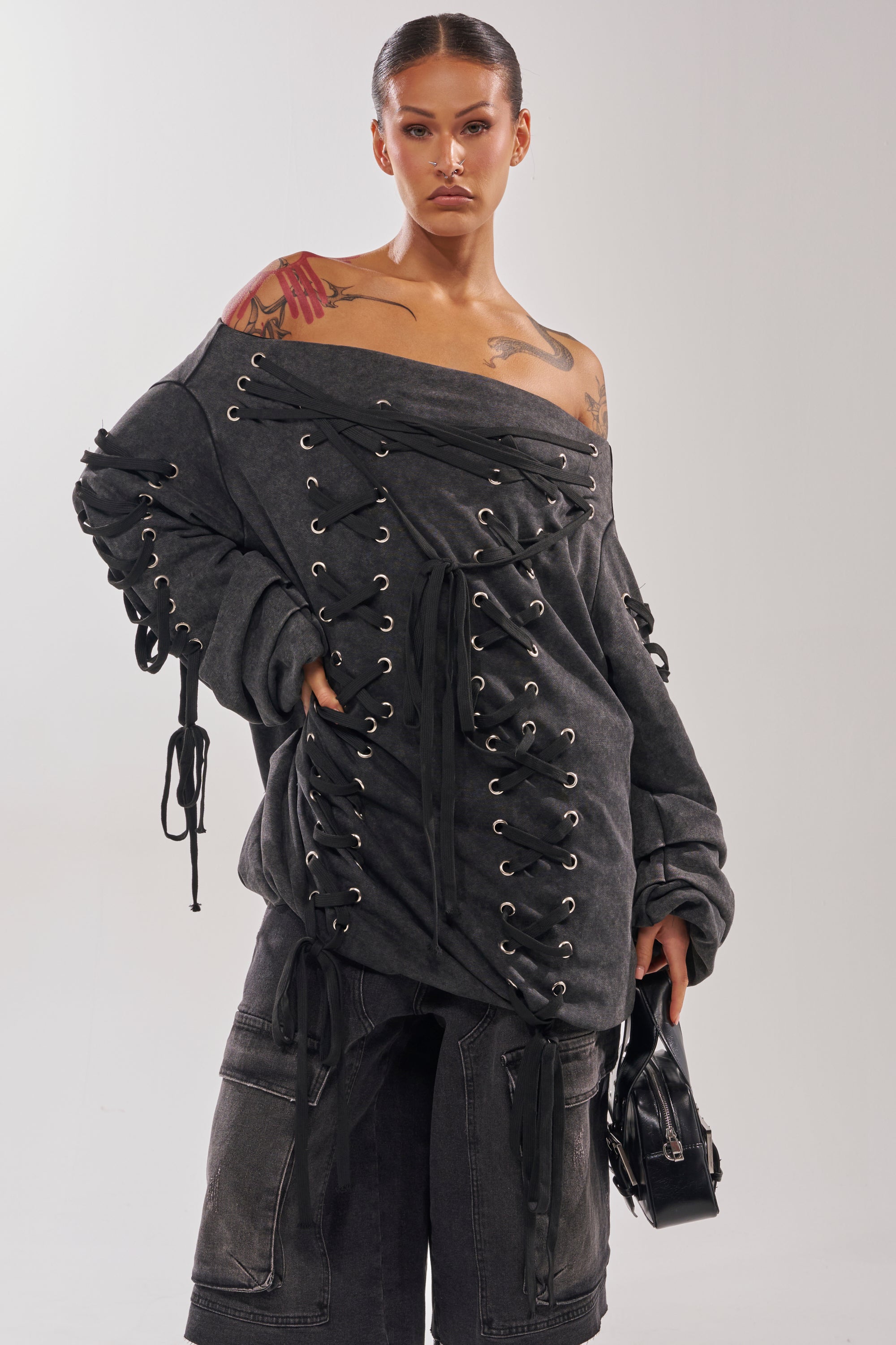 A person with slicked-back hair wears the LACED UP FOR GOOD MINI DRESS, an off-shoulder charcoal gray dress with lace-up detailing. They carry a small black bag and show tattoos on their shoulder and arm.