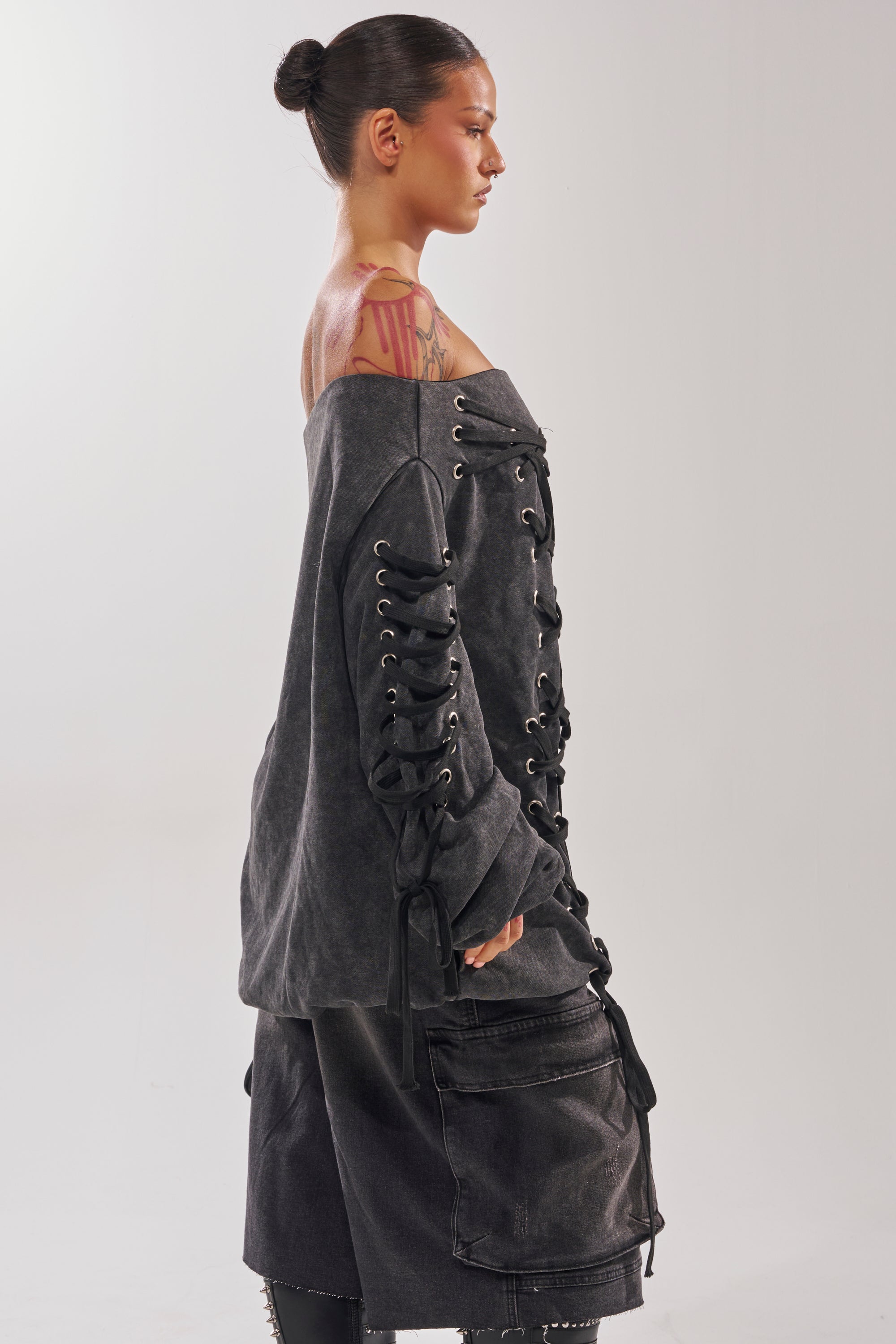 A person with a bun hairstyle and visible shoulder tattoos exudes street-chic style in the LACED UP FOR GOOD MINI DRESS, an off-the-shoulder, lace-up dark gray dress, against a plain light background.