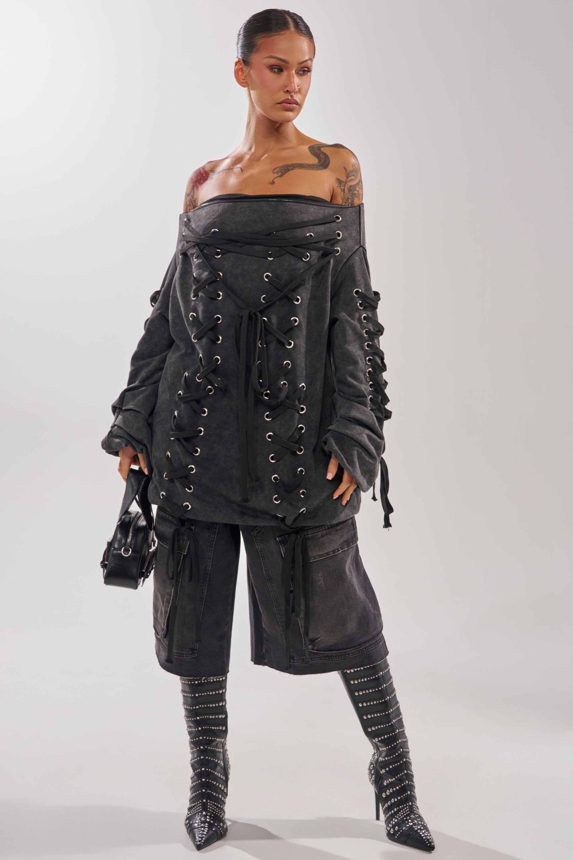 A woman in the LACED UP FOR GOOD MINI DRESS, an off-the-shoulder oversized charcoal gray piece with lace-up details, pairs it with knee-high studded boots and a small black bag. She sports slicked-back hair and shoulder tattoos.