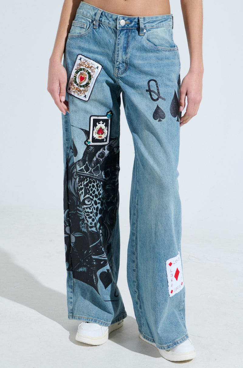 A person wears the SLEIGHT OF HAND RELAXED JEANS, featuring wide-leg high-rise design with playing card patches and a black leopard graphic on one leg, highlighting spade, club, heart, and diamond symbols. They pose against a plain backdrop.