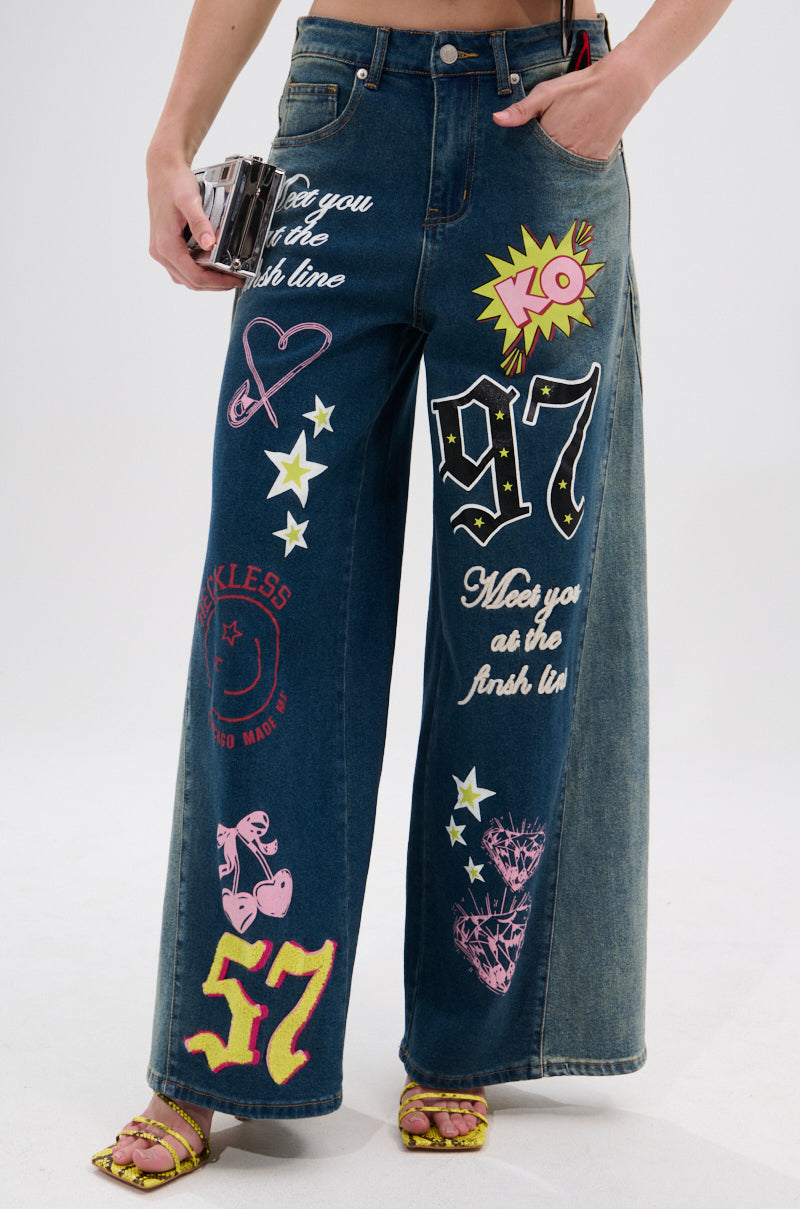 Wearing the TKO GRAPHIC WIDE LEG DENIM JEAN with high-rise fit, graphic patches, "97" and "57," stars, and phrases like “Meet you at the finish line,” paired with a small clutch and yellow sandals.