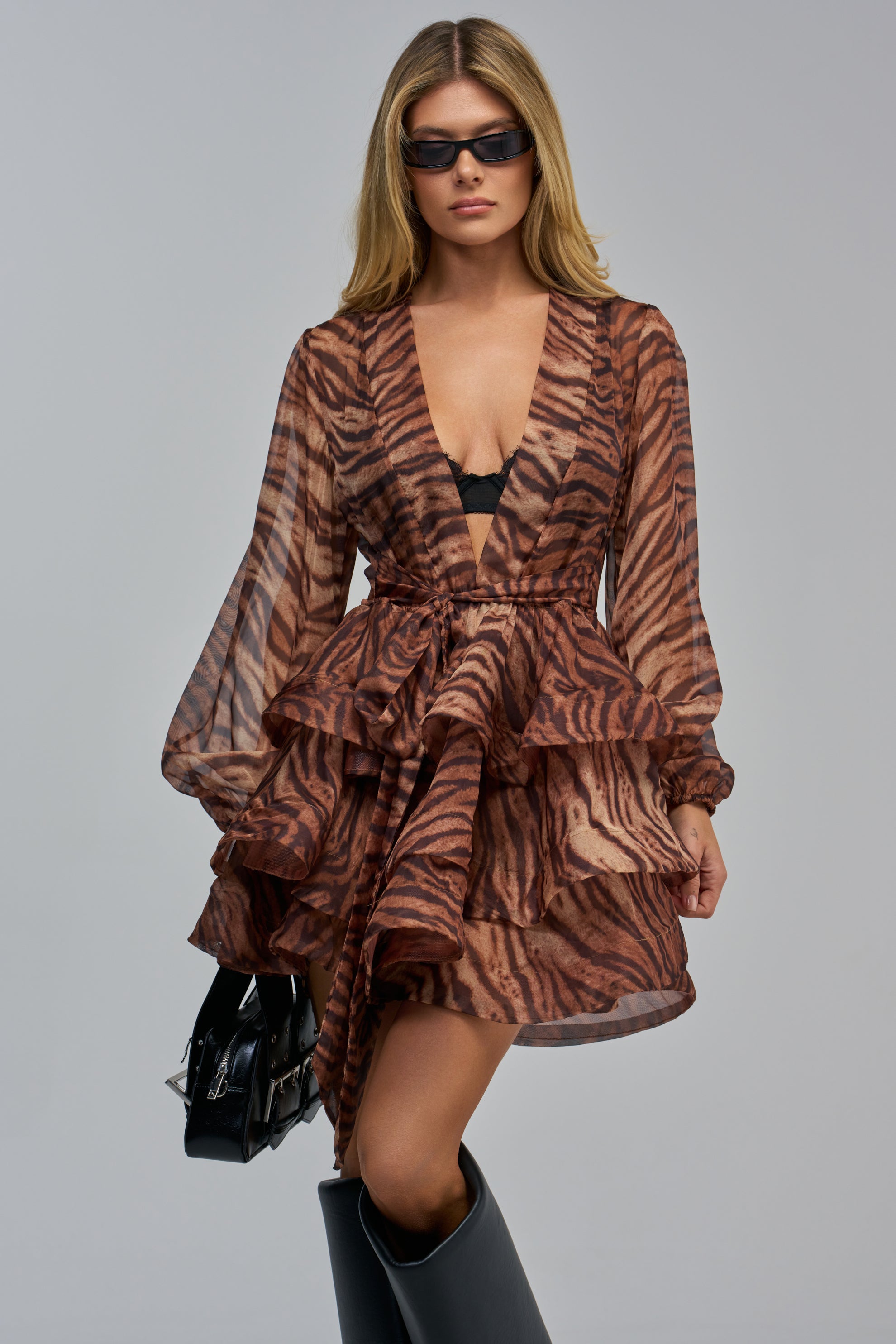 A model wearing a SEA BREEZE RUFFLE MINI DRESS IN TIGER, paired with black sunglasses, black knee-high boots, and a black handbag.