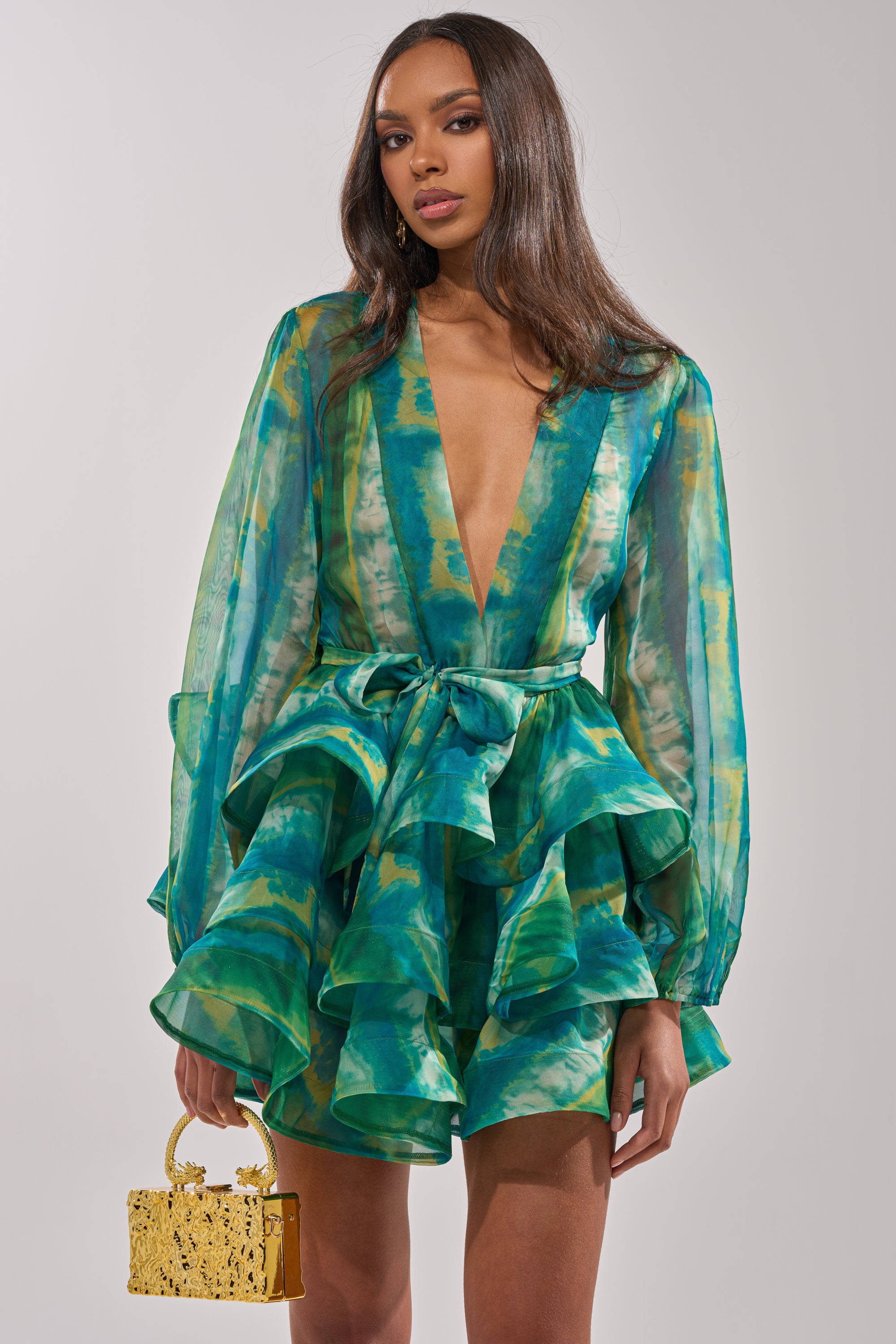 A woman in the SEA BREEZE RUFFLE MINI DRESS, a vibrant green and blue chiffon mini with deep V-neckline and long sleeves, holds a small gold handbag against a plain light background—the perfect chic resort wear.