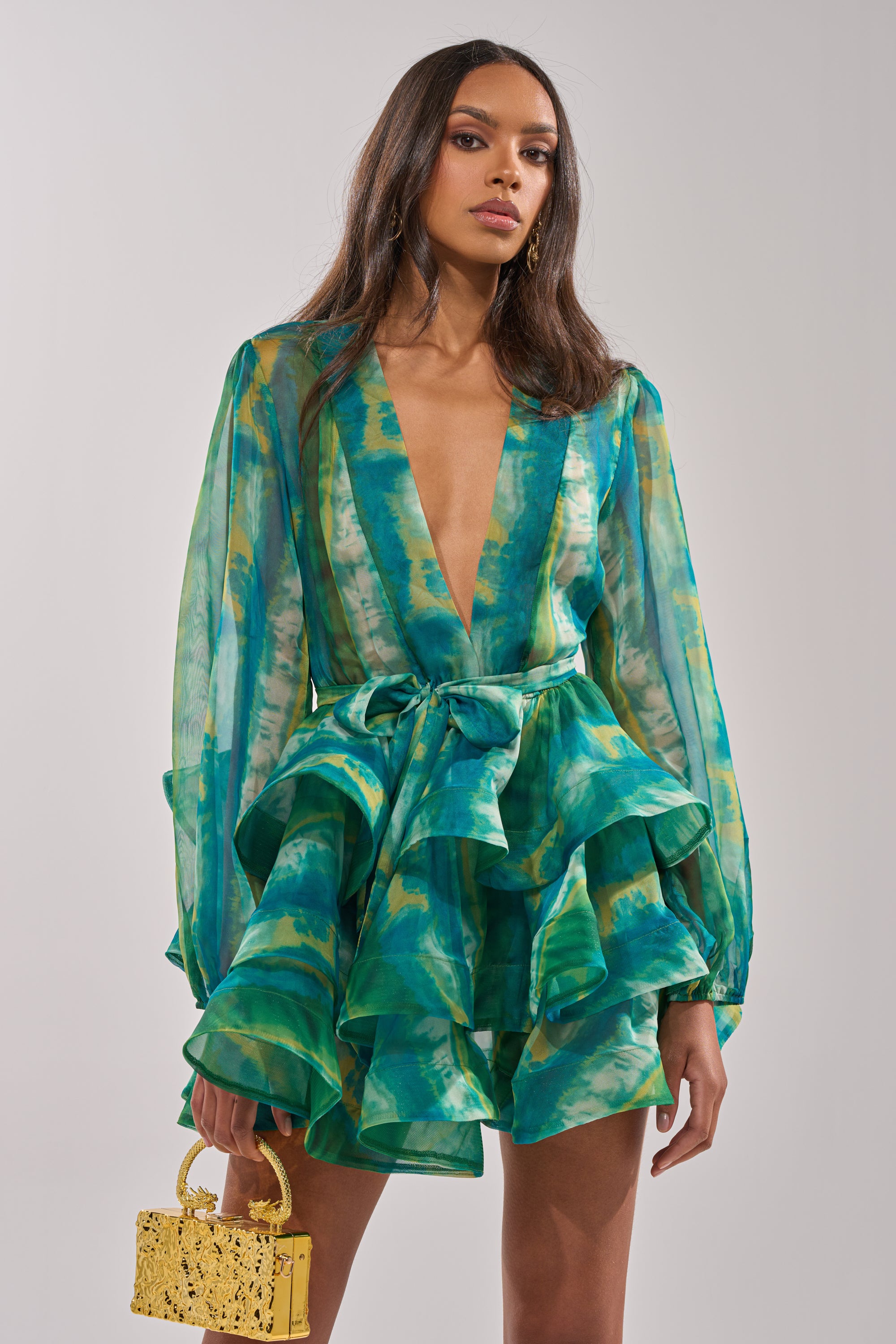 A woman with long brown hair and a confident expression models the SEA BREEZE RUFFLE MINI DRESS, featuring a deep V-neck, long sleeves, and green-blue tie-dye chiffon ruffles. She stands against a plain background holding a small yellow handbag.