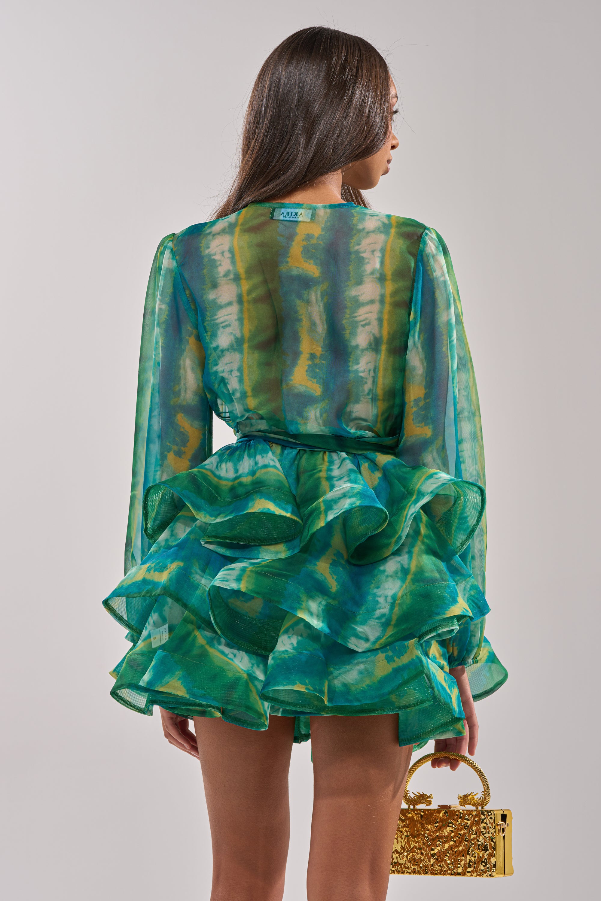 A woman with long brown hair models the SEA BREEZE RUFFLE MINI DRESS—a sheer green and yellow ruffled chiffon mini—facing away and holding a small gold handbag against a plain, light background.