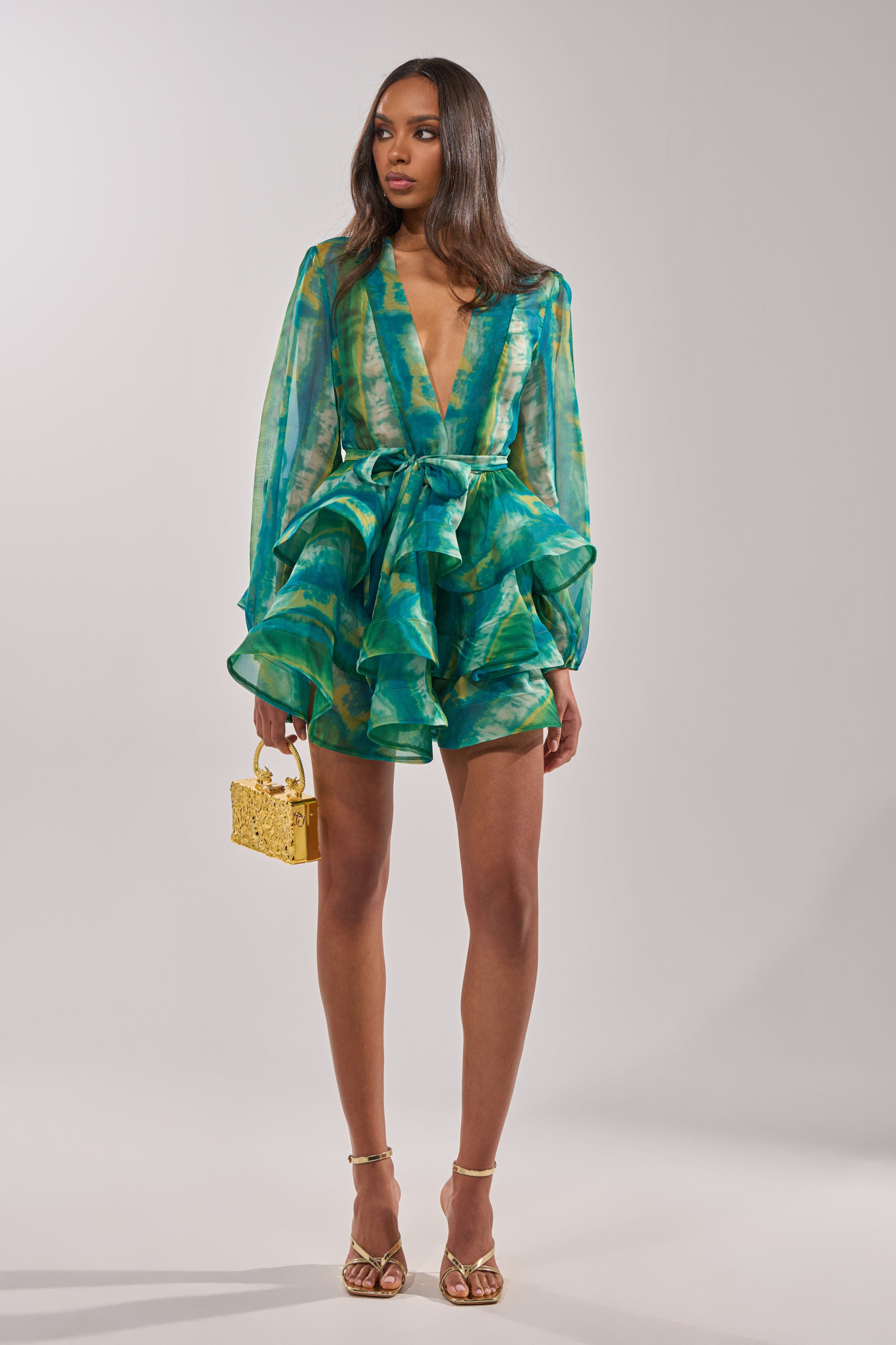 A woman stands against a plain backdrop in the SEA BREEZE RUFFLE MINI DRESS—a green and blue patterned romper with a deep v-neck, sheer long sleeves, ruffled shorts, tie waist belt—styled with gold heels and a small gold handbag.