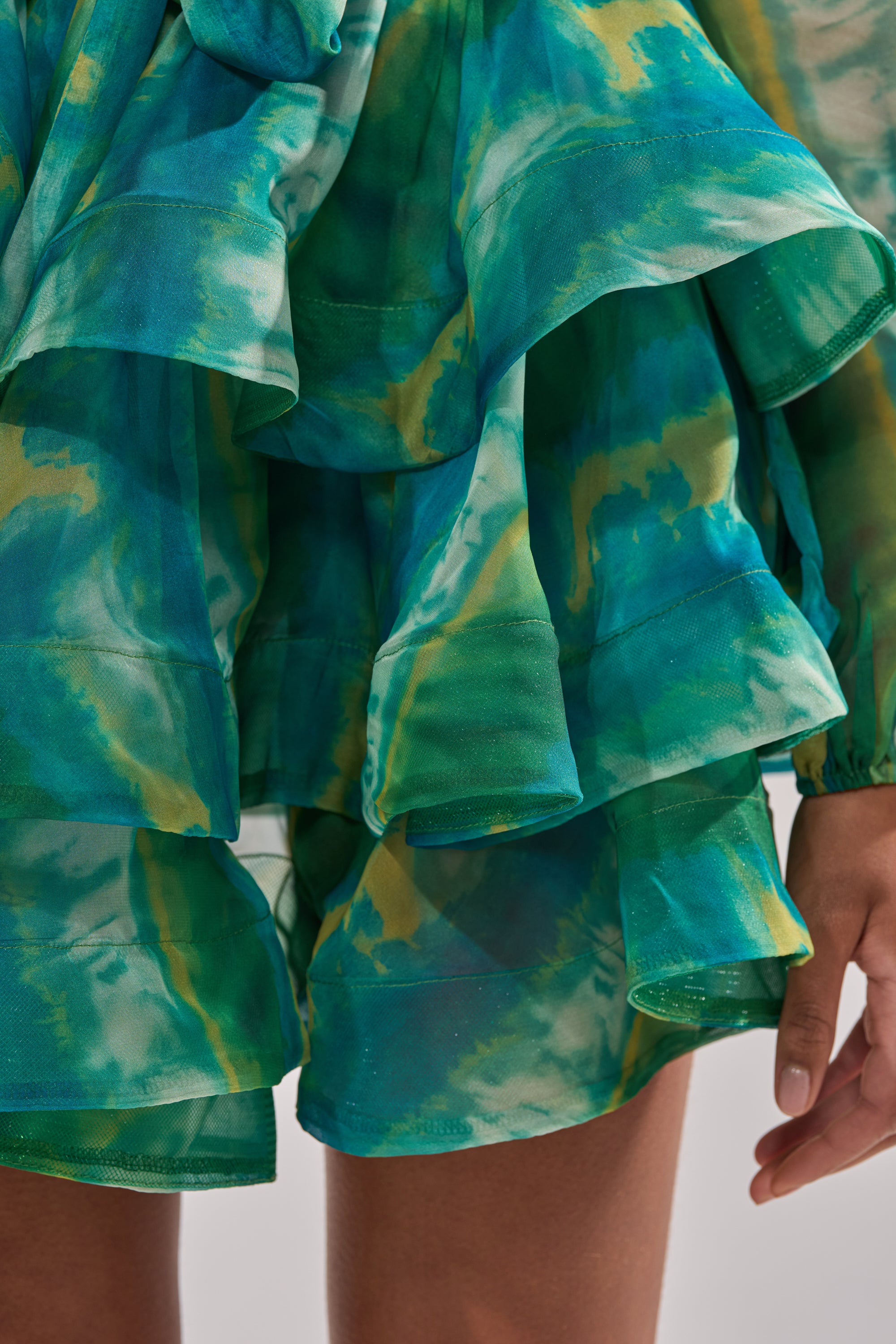 A close-up shows a person wearing the SEA BREEZE RUFFLE MINI DRESS, featuring green, blue, and yellow watercolor patterns. The layered chiffon design reveals part of their hand and leg, highlighting its elegant resort style.