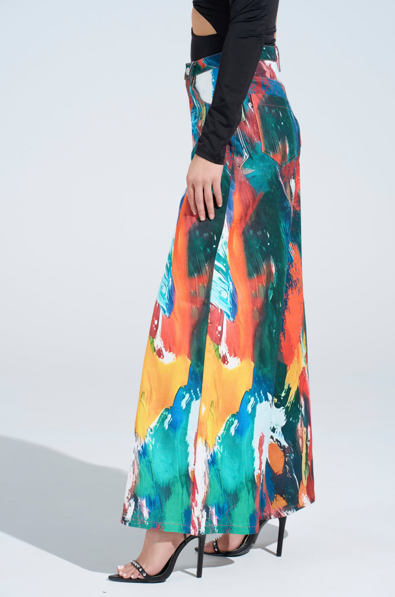 Clad in the Creative Freedom Printed Denim, a person with an artistic vibe stands sideways, pairing the colorful maxi skirt with a black top and sleek high heels. The simple light backdrop lets their creative flair shine.