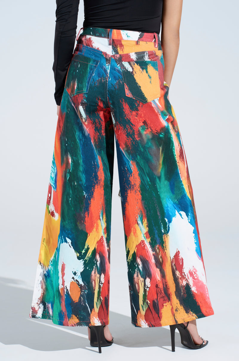 In a CREATIVE FREEDOM PRINTED DENIM ensemble of a black one-sleeve top and abstract-patterned wide-leg pants, a person stands artistically with their back to the camera, paired effortlessly with black high heels against a plain backdrop.