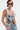 A person with short black hair and a shoulder tattoo wears sunglasses, a silver choker, and the OUT OF MY LEAGUE DENIM HALTER TOP featuring graphic prints and colorful florals, paired with matching denim pants.