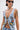 A person wearing sunglasses, a silver choker, and the OUT OF MY LEAGUE DENIM HALTER TOP with colorful floral and animal embroidery, plus visible arm and chest tattoos, stands against a plain white background.