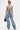 Wearing the OUT OF MY LEAGUE DENIM HALTER TOP with matching flared light blue denim pants, both featuring colorful floral embroidery and bold black prints, the person styles short hair, sunglasses, a statement necklace, and a black handbag.
